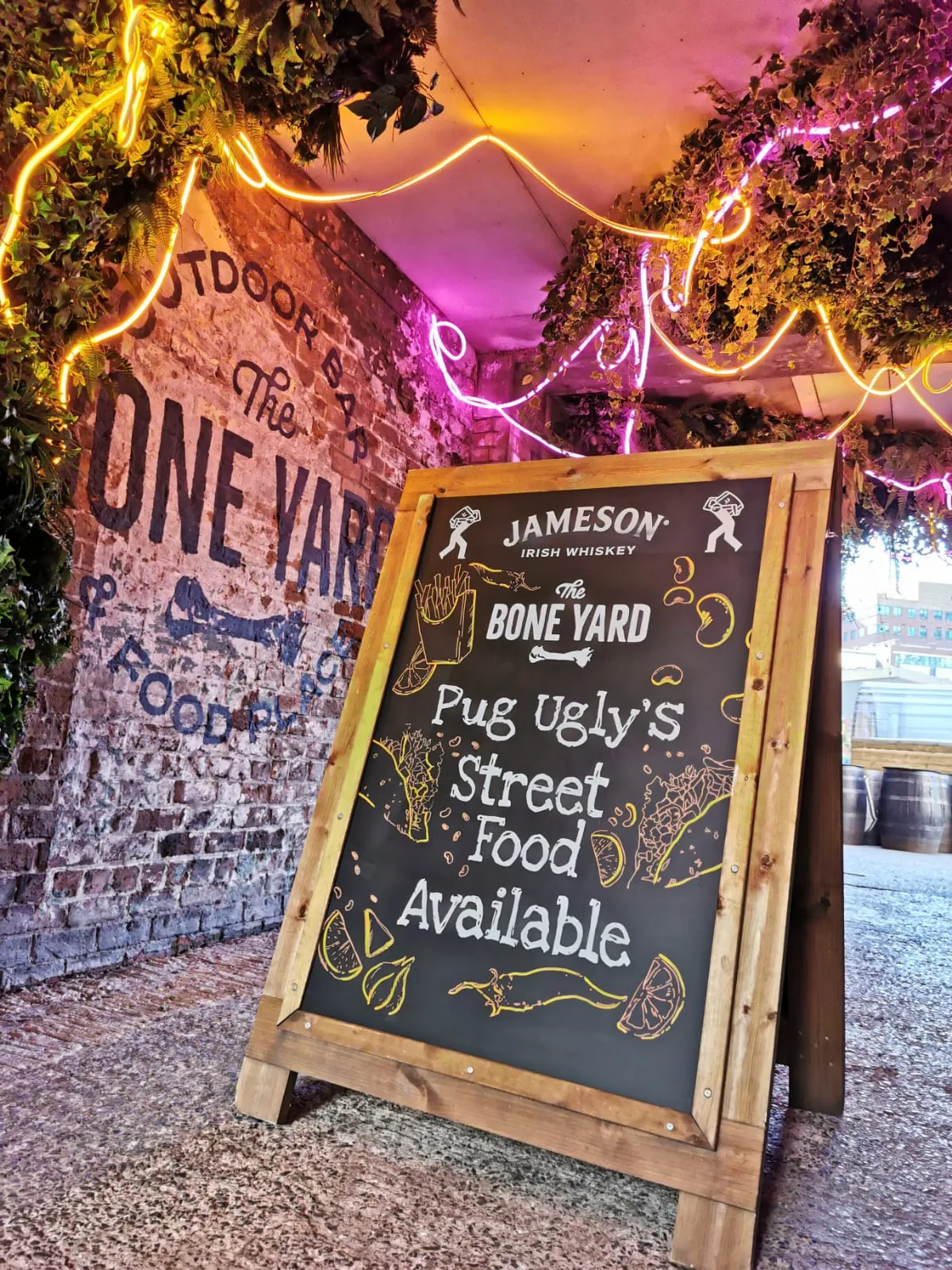 A wooden A-board under pink and yellow neon at The Bone Yard, its black panel lettered “Pug Ugly’s Street Food Available” with Jameson branding and little food illustrations.