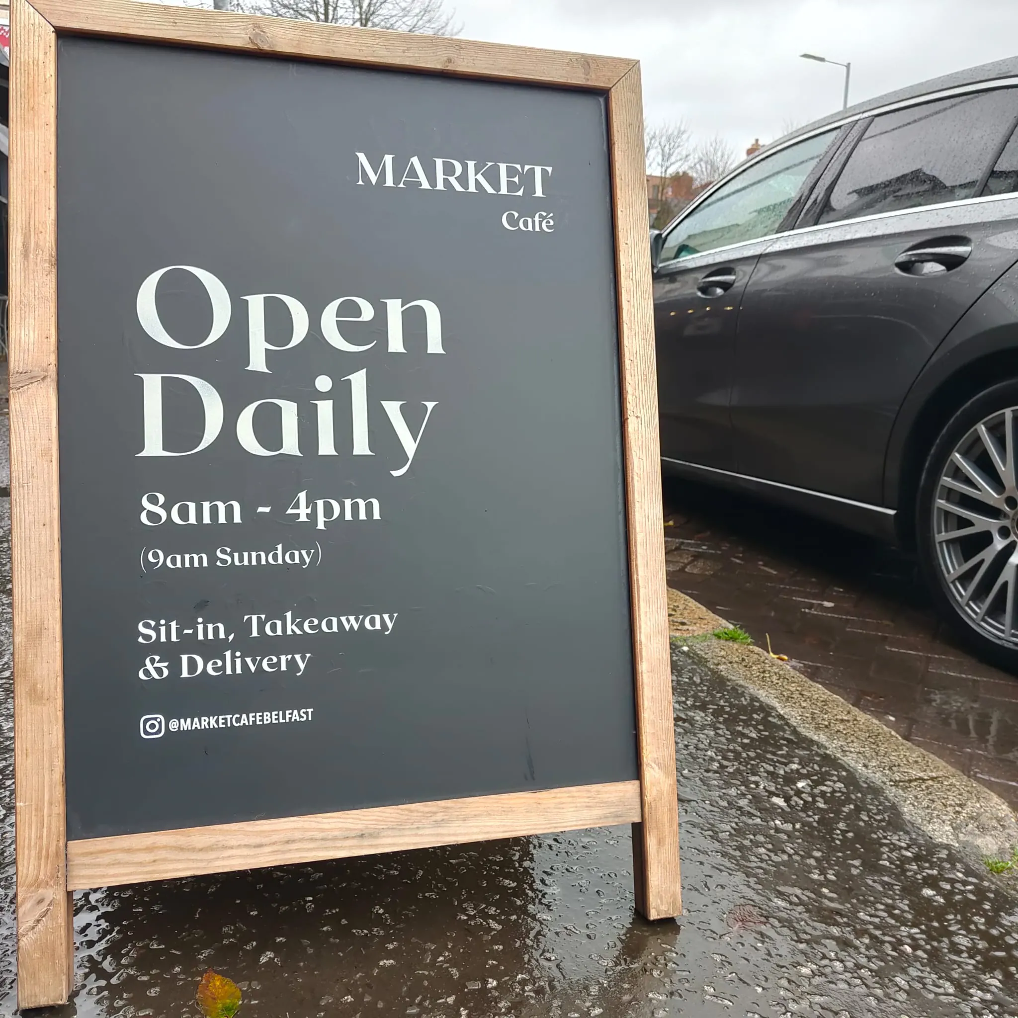 A wooden A-board for Market Café reading “Open daily, 8am – 4pm” in white serif lettering on a black panel, standing on a wet pavement.