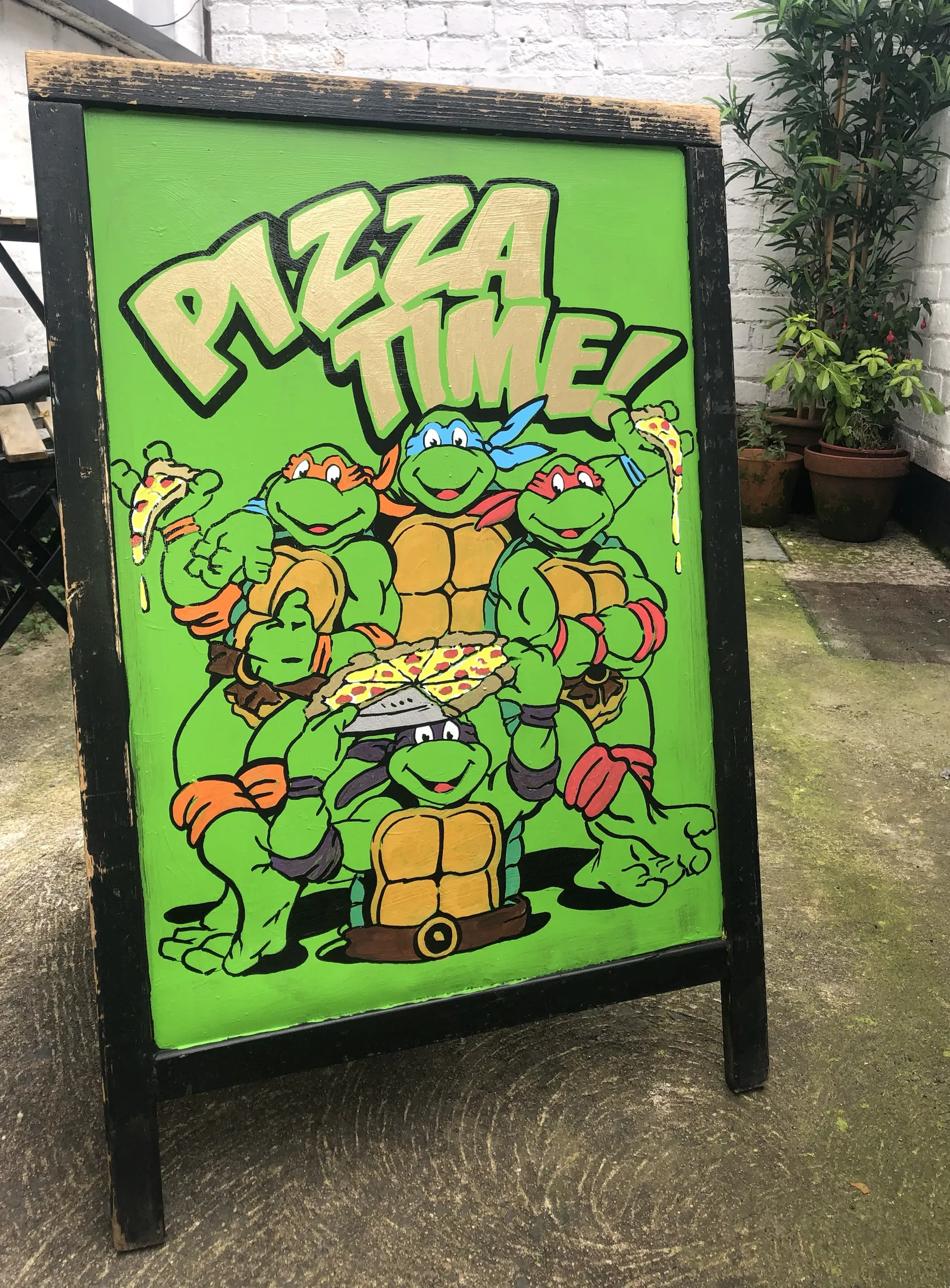 A green A-board hand-painted with the Teenage Mutant Ninja Turtles sharing a pizza under the words “Pizza Time!”.