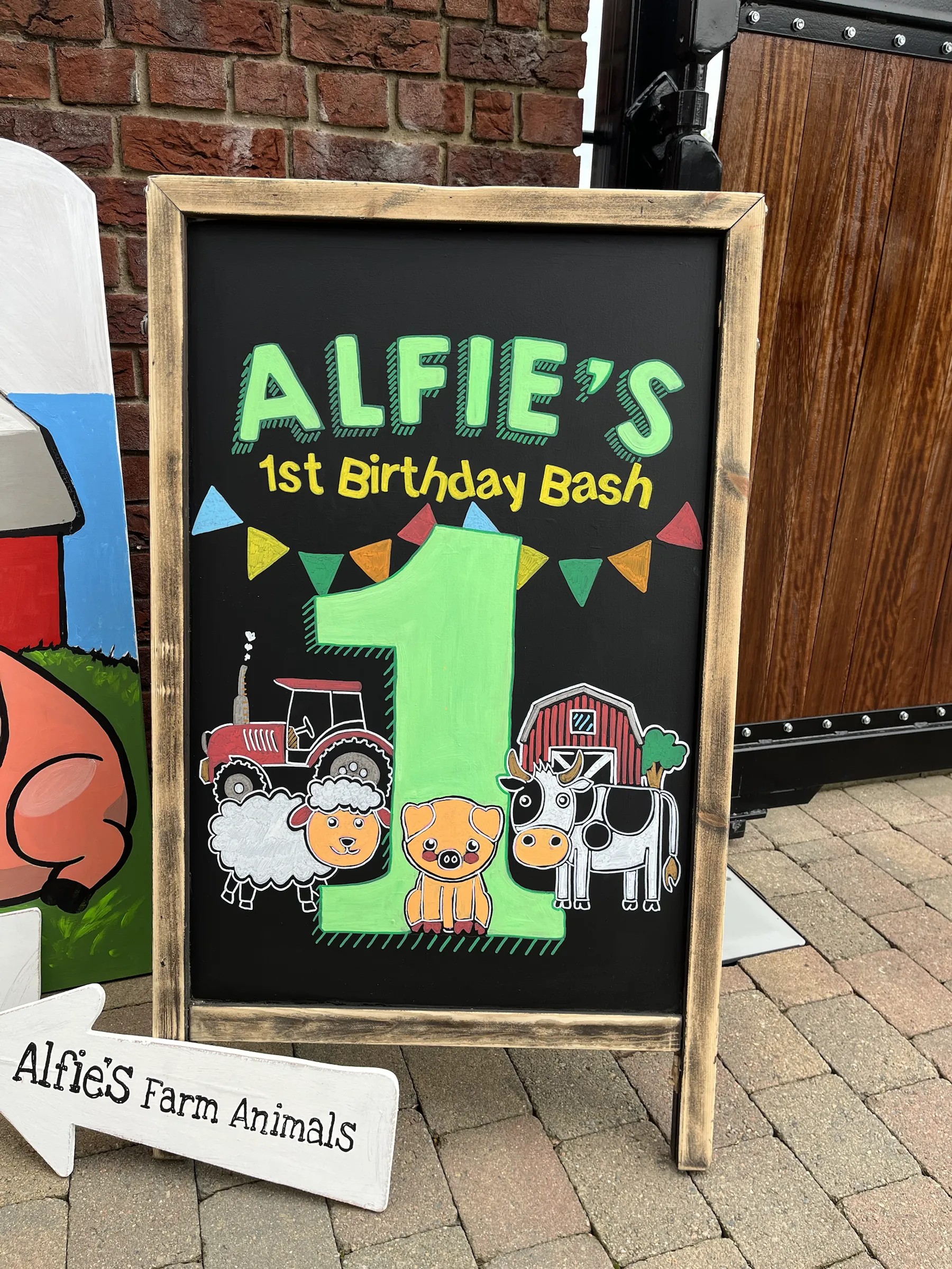 A wooden A-board for Alfie’s 1st Birthday Bash, painted with bunting, a big green number one and a tractor, barn and farm animals.