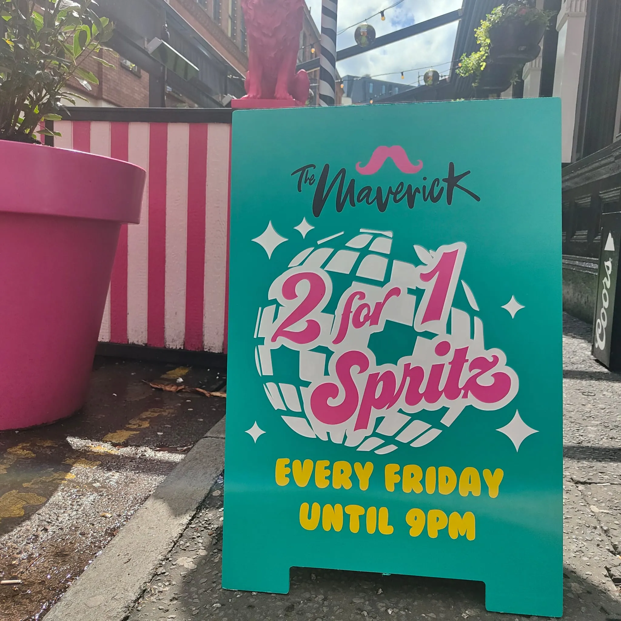 A teal A-board for The Maverick advertising “2 for 1 Spritz, every Friday until 9pm” over a pink disco ball, beside striped planters.
