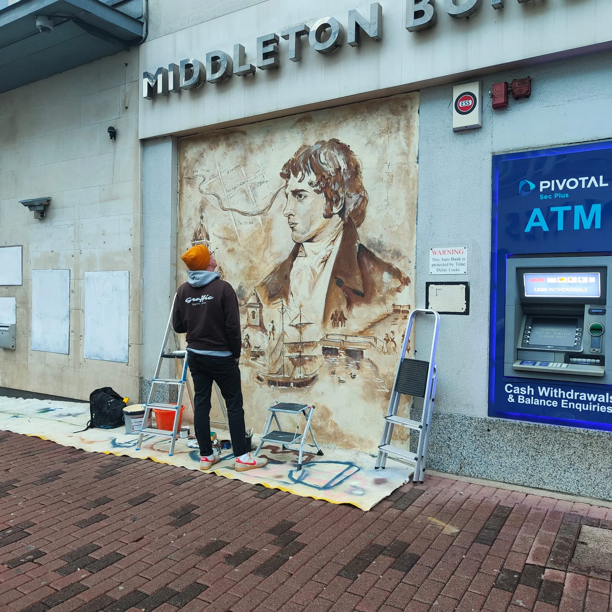 A painter in an orange beanie at work on the sepia High Street mural, beside an ATM.
