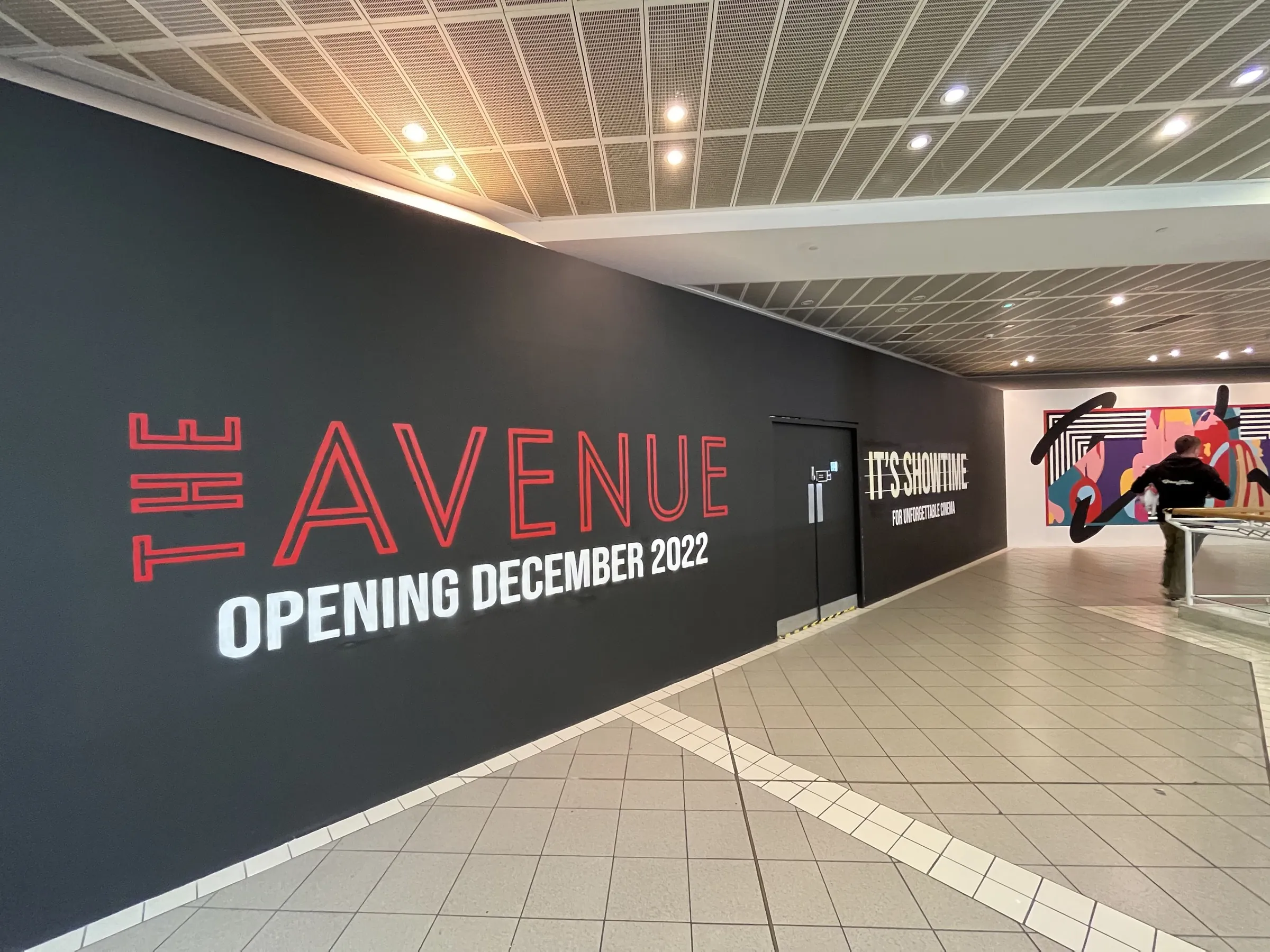 A long black hoarding lettered “The Avenue, opening December 2022” and “It’s Showtime”, the abstract mural visible at the far end of the walkway.