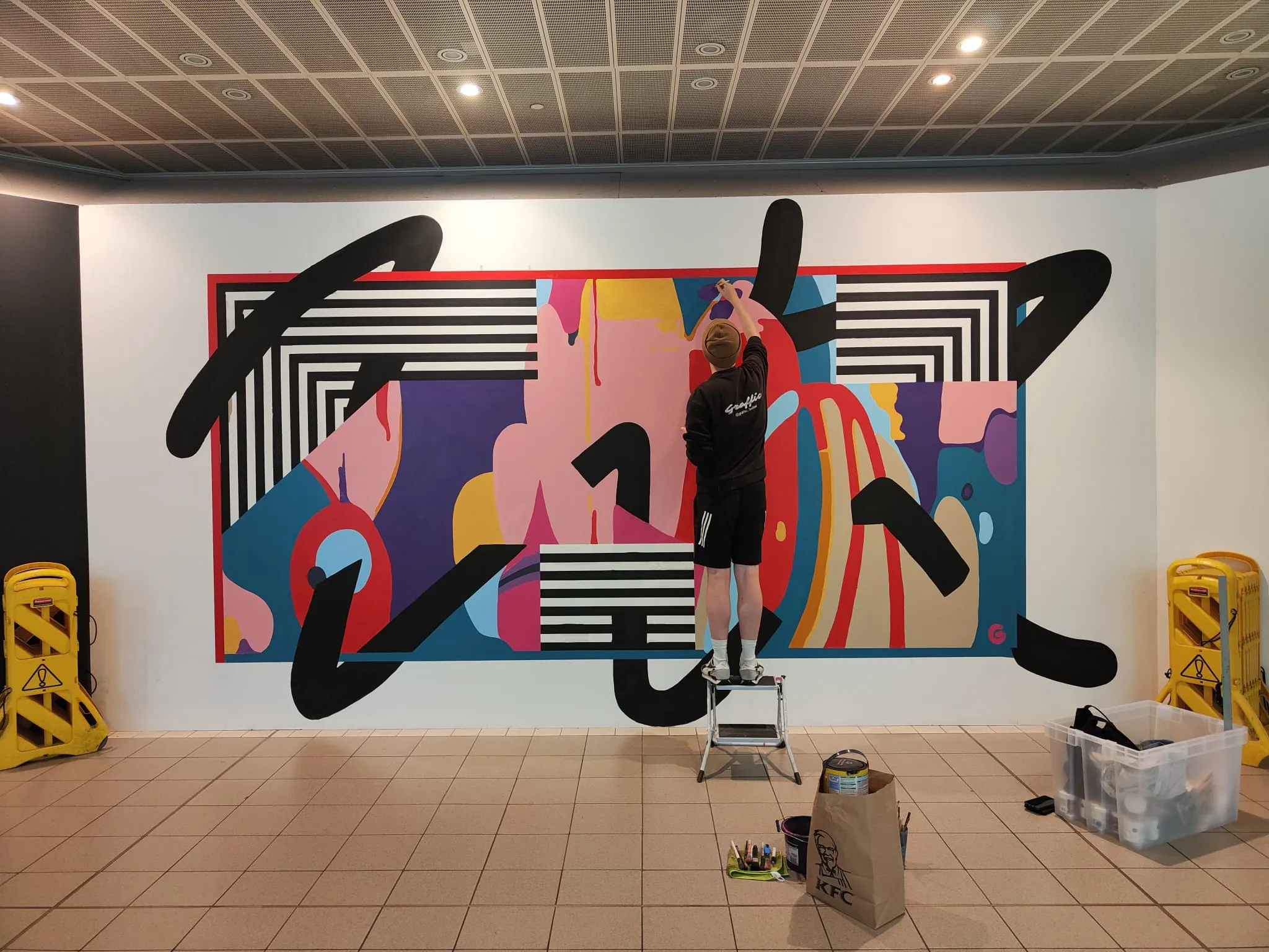 A Graffic painter on a step stool filling in the abstract mural’s pink and red shapes, yellow barriers and paint kit on the tiles below.