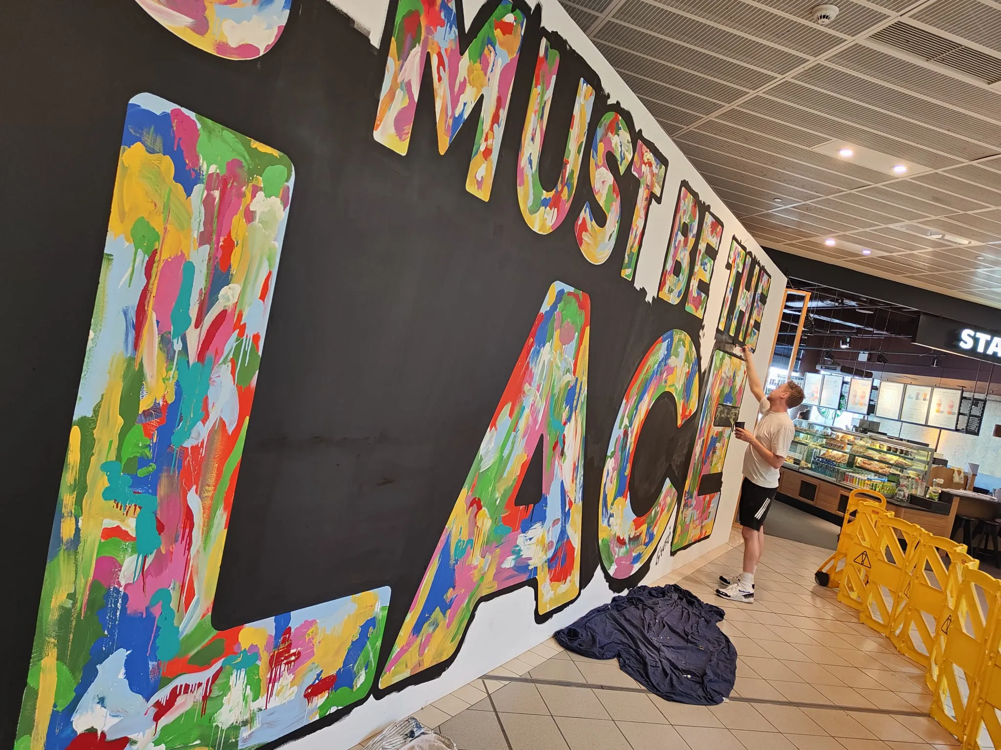 Giant multicoloured lettering on a black hoarding, a painter working at its far end behind yellow barriers.