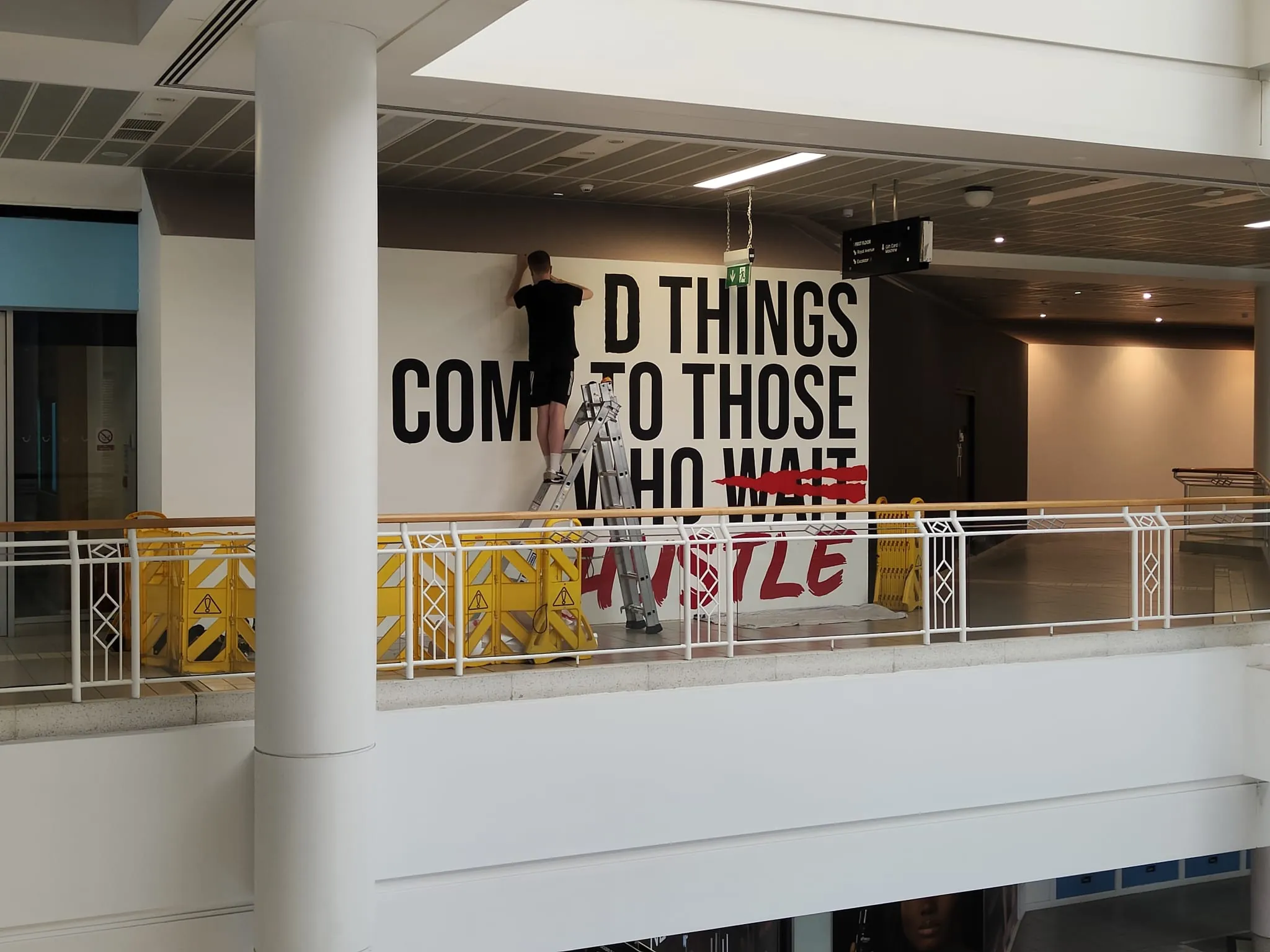 A painter up a ladder on the first-floor gallery, lettering “Good things come to those who wait” on a white hoarding.