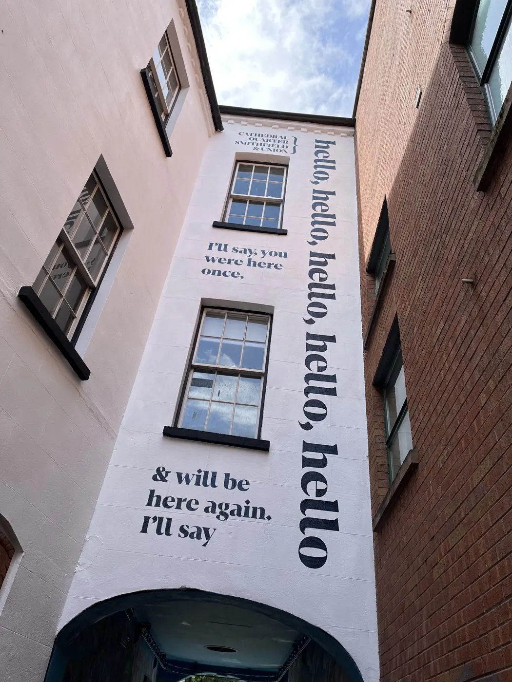 A mural commissioned by Cathedral Quarter, Smithfield and Union in Belfast. It is large, black text painted onto a white wall, around windows. The text reads 'I’ll say, you were here once, and will be here again. I’ll say hello, hello, hello, hello, hello'.