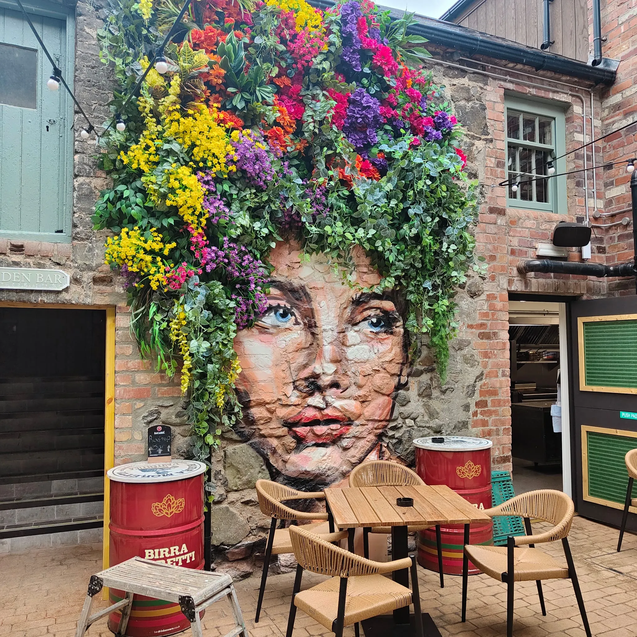 A woman’s face painted on the stone wall beneath a living crown of real flowers, above the courtyard tables.