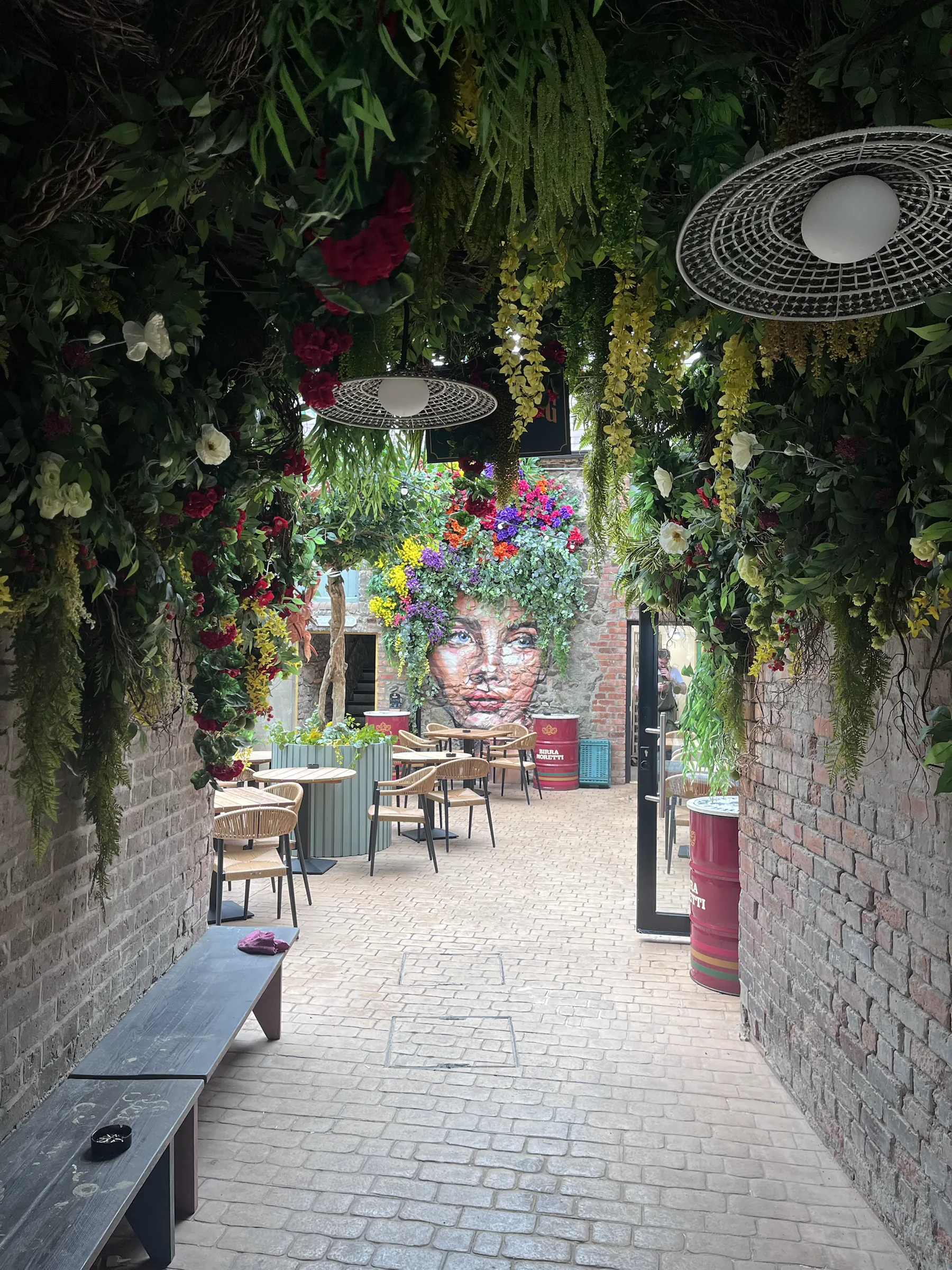 Eden’s flower-filled alley under hanging greenery, with the painted face mural at its far end.
