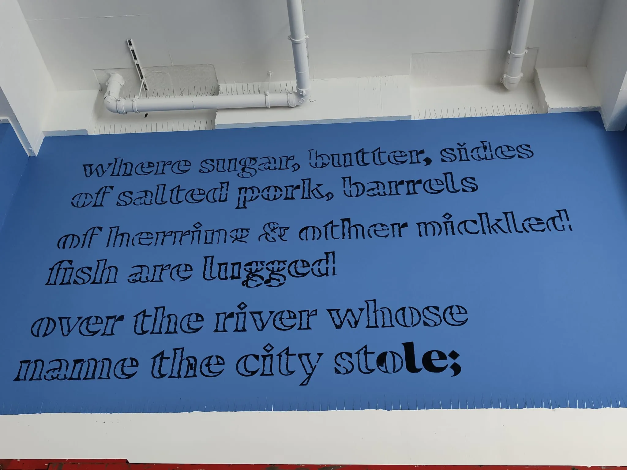 Lines of the poem lettered in black outline on a blue wall: “where sugar, butter, sides of salted pork, barrels of herring & other pickled fish are lugged over the river whose name the city stole;”.