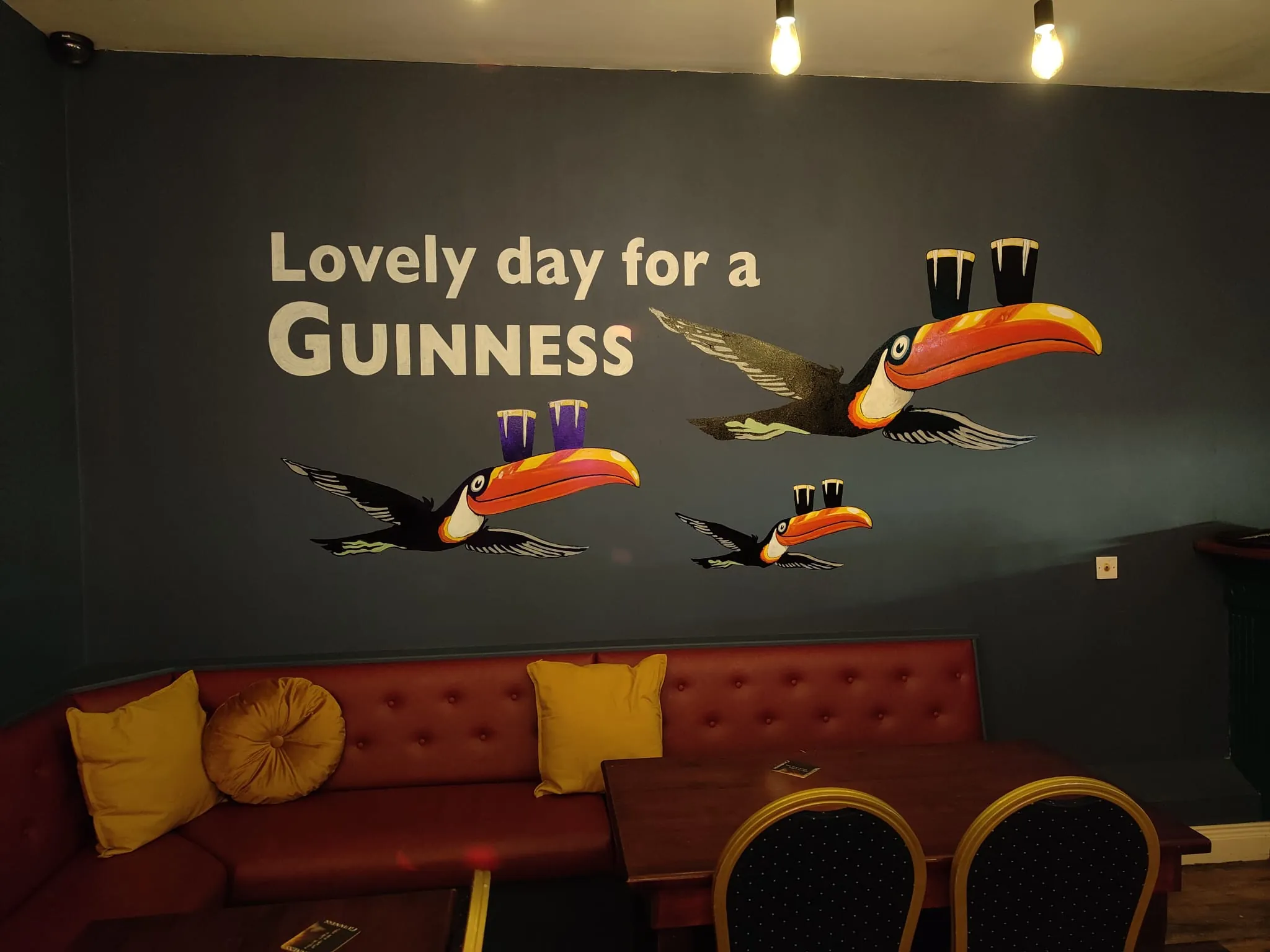 A navy wall at the Fiddlers painted with three Guinness toucans under “Lovely day for a Guinness”, above red leather booths.