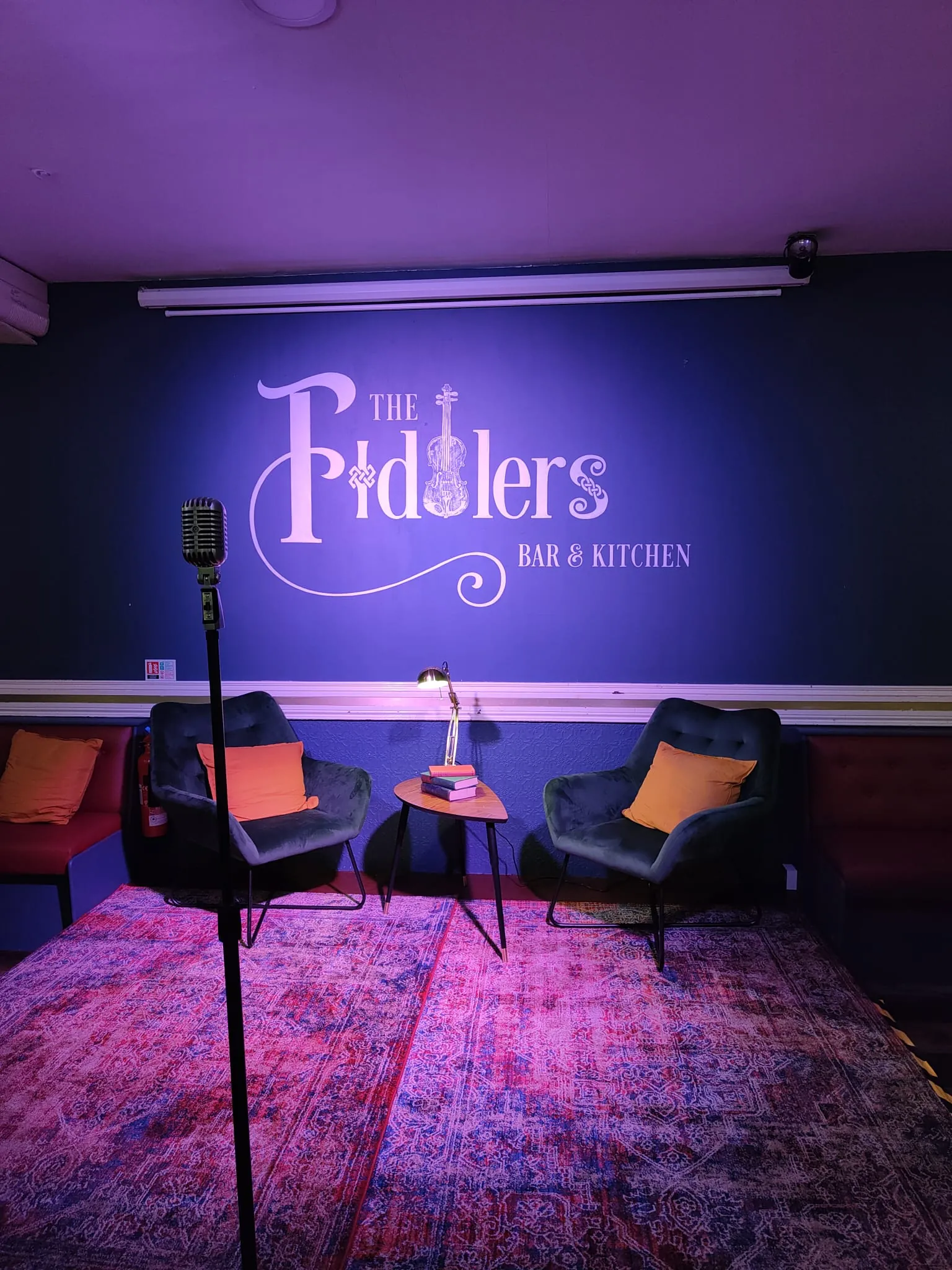 The Fiddlers logo painted on a blue-lit stage wall behind a microphone stand and two armchairs.