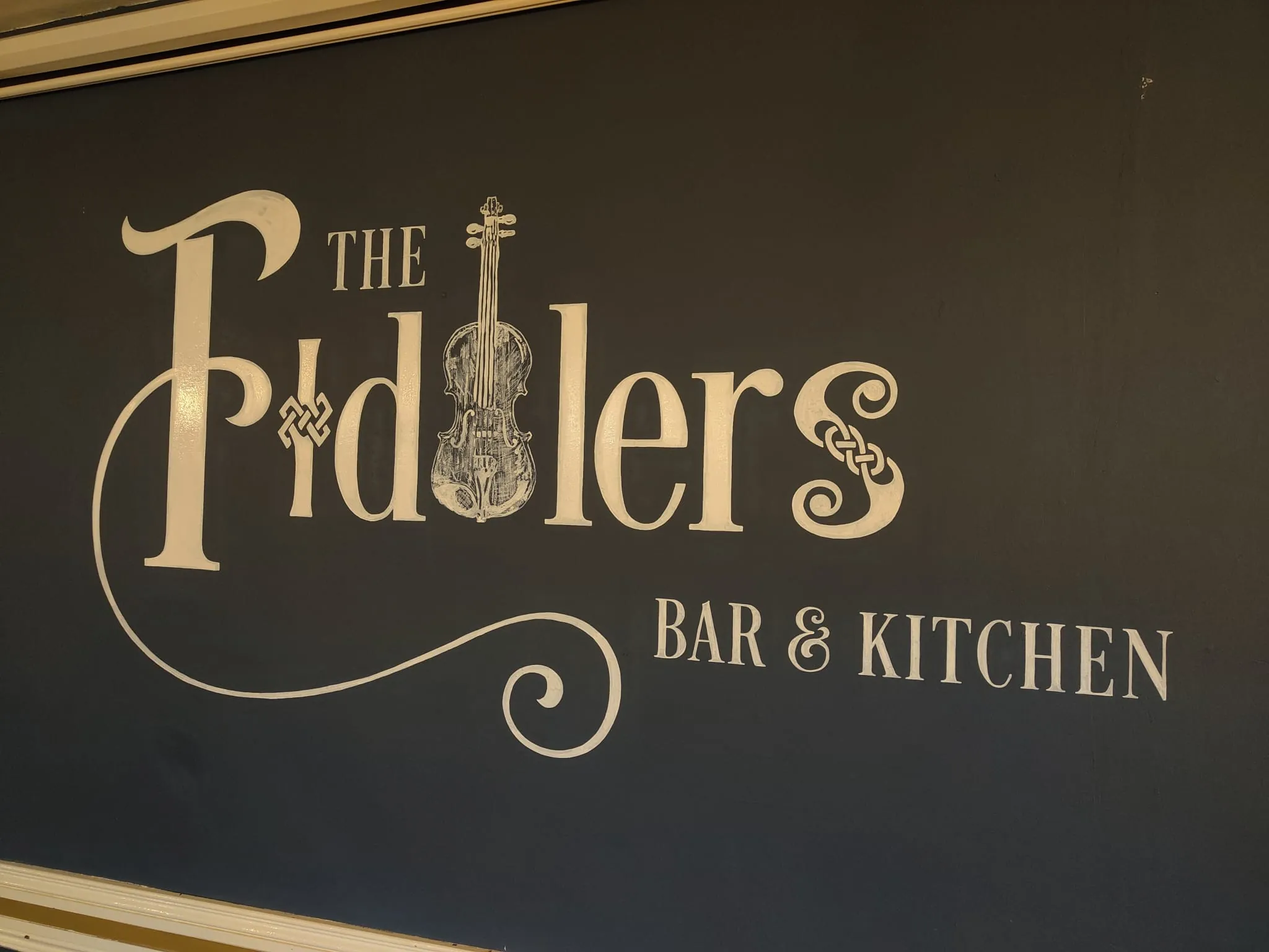 The Fiddlers logo, a fiddle forming its “dd”, painted in cream above “Bar & Kitchen” on a dark wall.