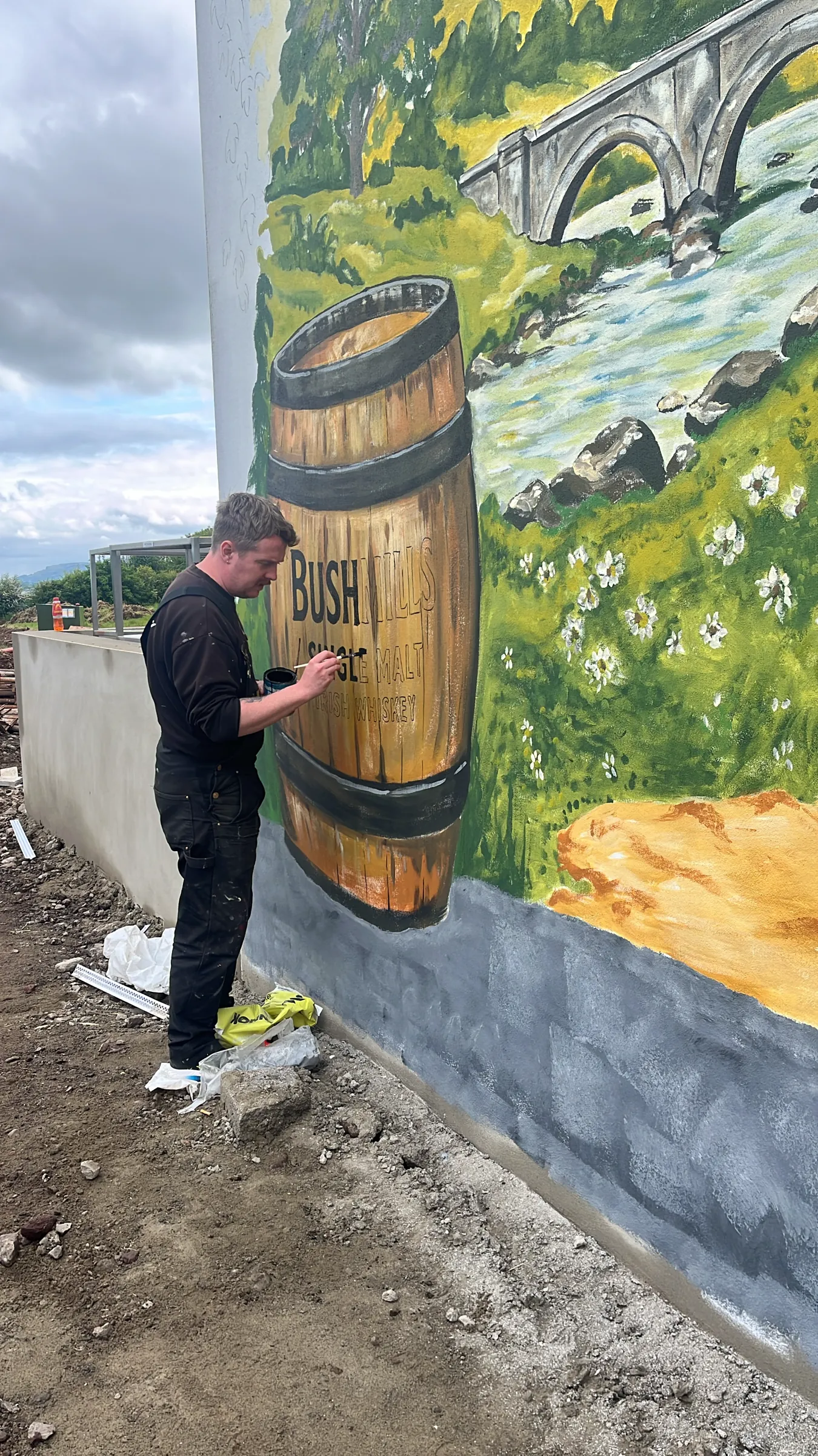 A painter at work on the Bushmills barrel, with a stone bridge and river painted above.