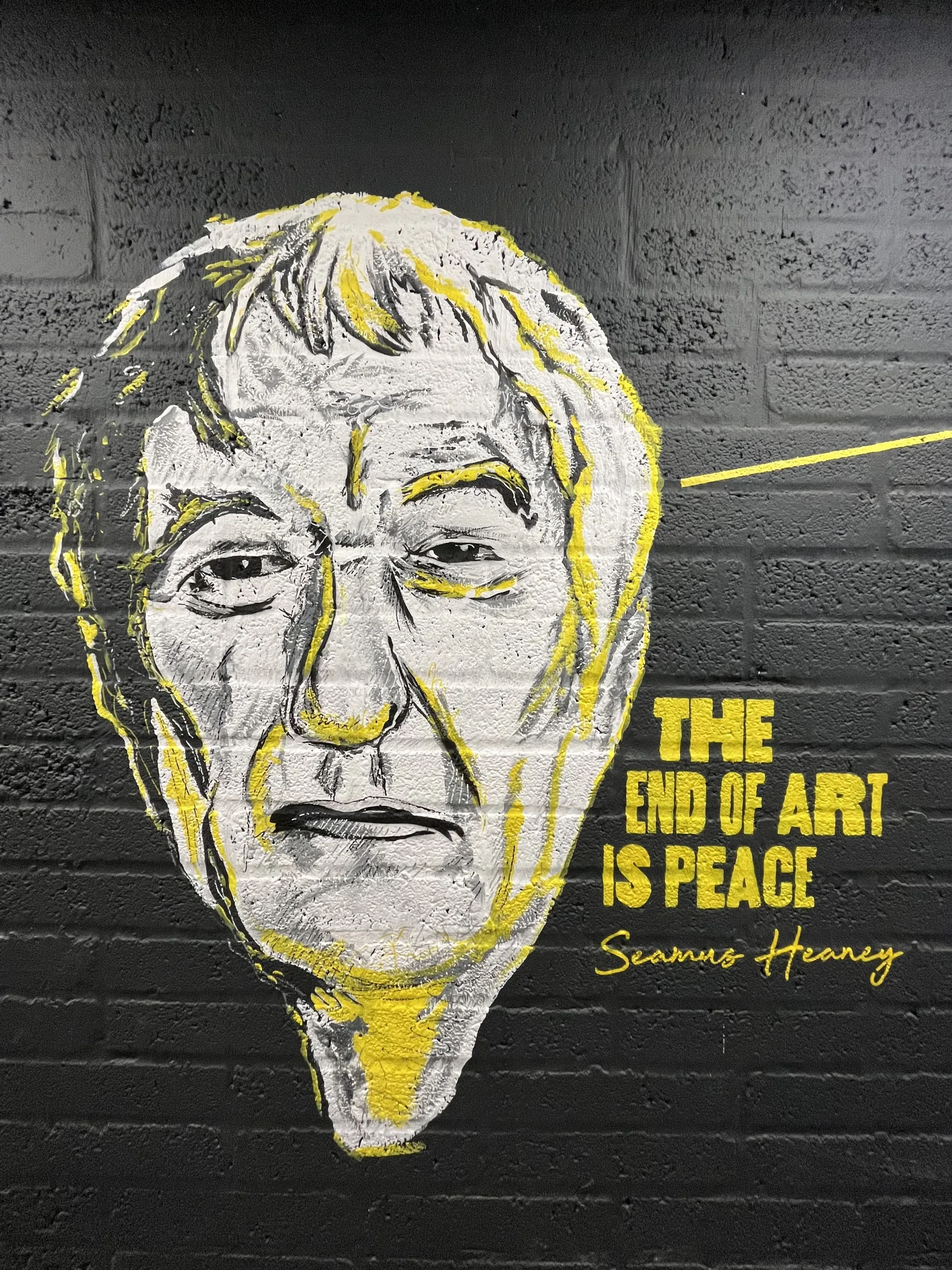A yellow and white portrait of Seamus Heaney captioned “The end of art is peace” on black brick.
