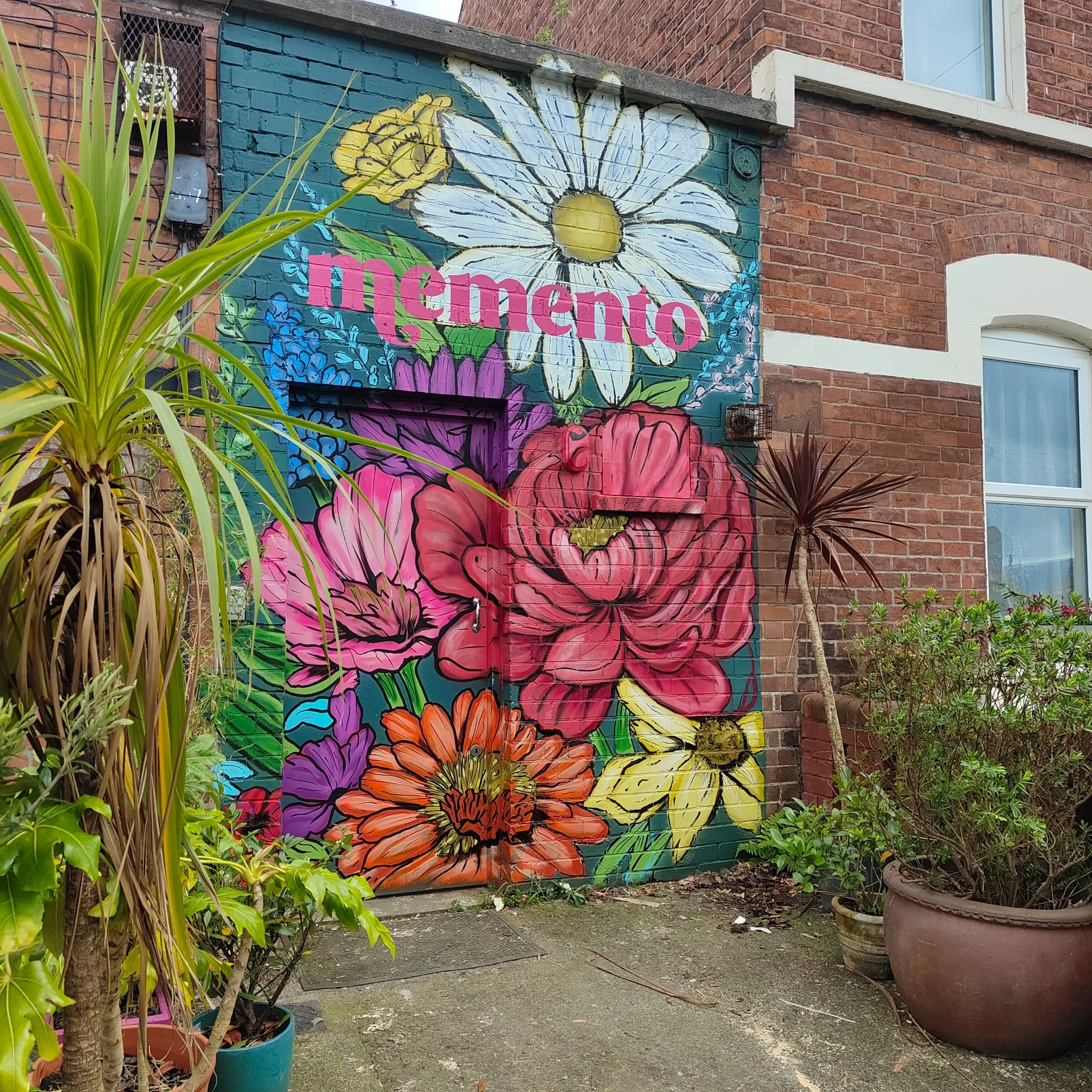 A garden wall painted with giant daisies, peonies and gerberas around the Memento name in pink.