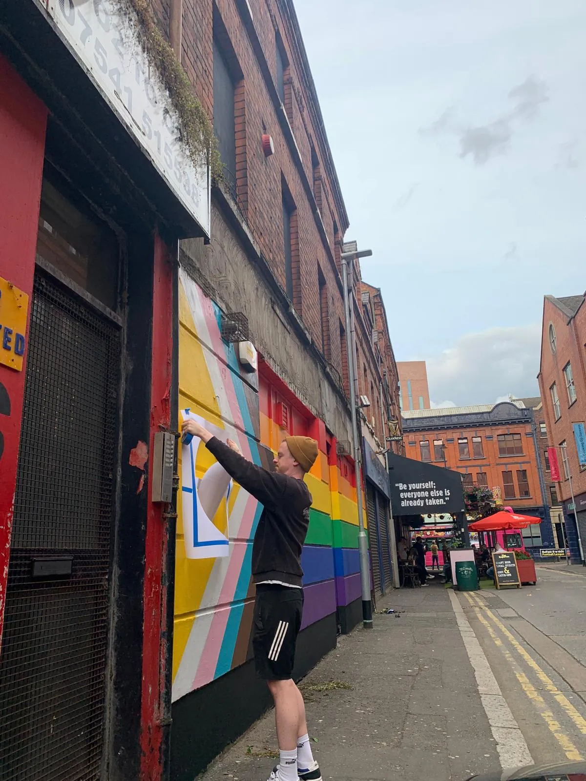 A painter in a beanie reaching up to paint a pride mural of rainbow and chevron stripes down the side of a red-brick building on a narrow street.