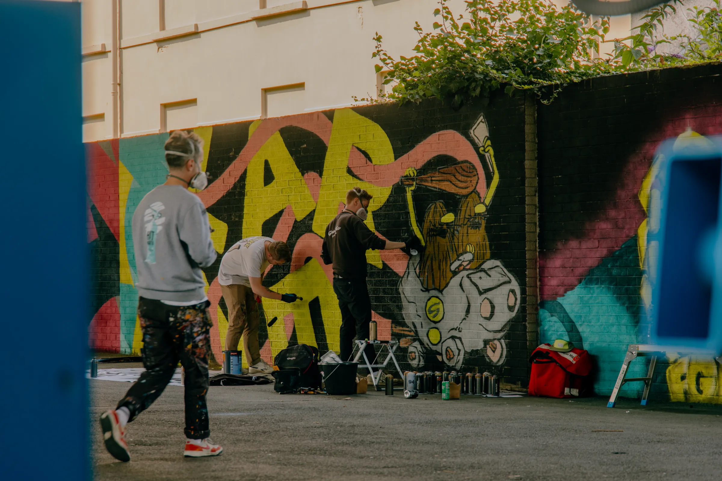 Painters at work on a long wall mural of colourful lettering and a cartoon character, with spray cans and kit laid out at their feet.