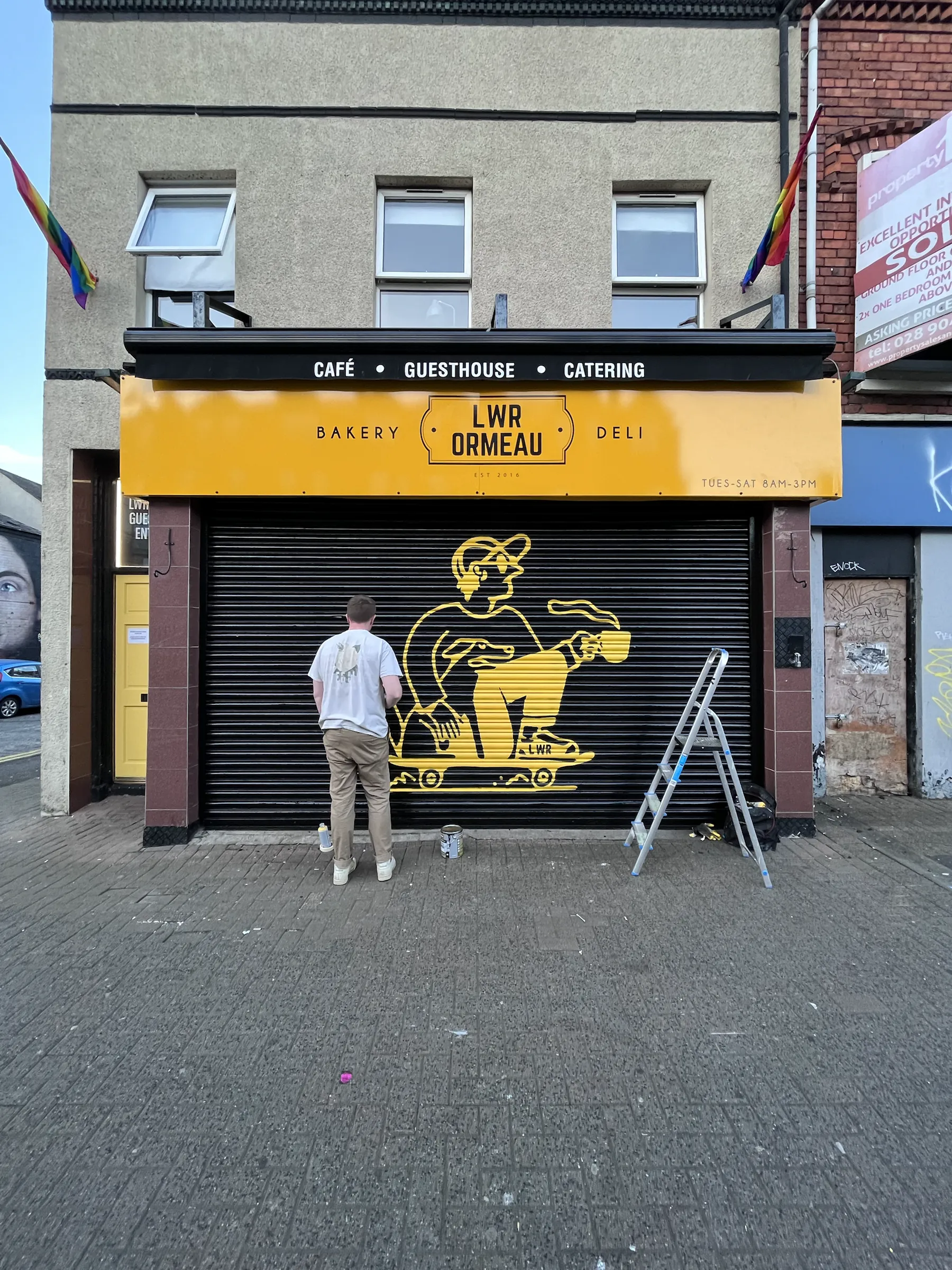 A painter at work on the shutter of Lwr Ormeau, painting a yellow line illustration of a figure on a skateboard carrying food, a stepladder alongside.
