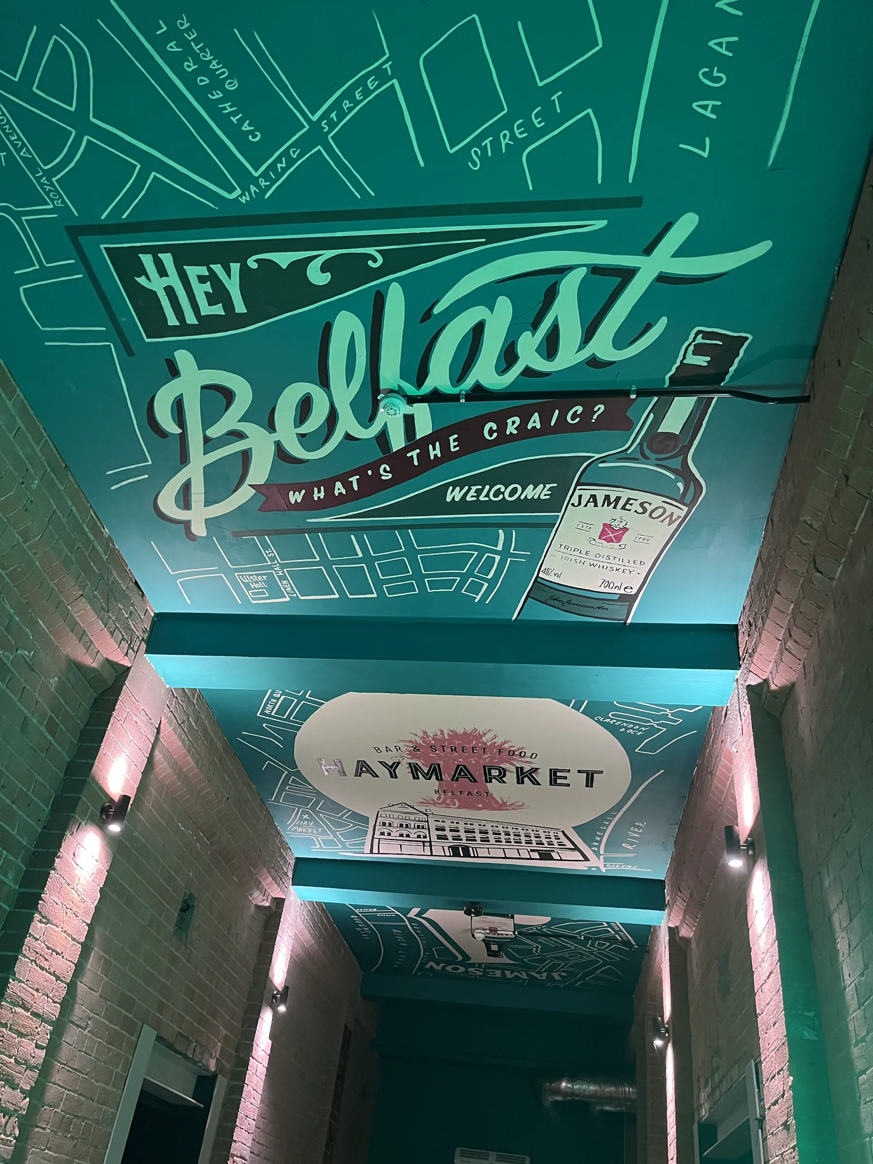 A teal-lit passageway ceiling painted with “Hey Belfast, what’s the craic? Welcome” over a line-drawn street map, with a Jameson bottle and the Haymarket name further along.