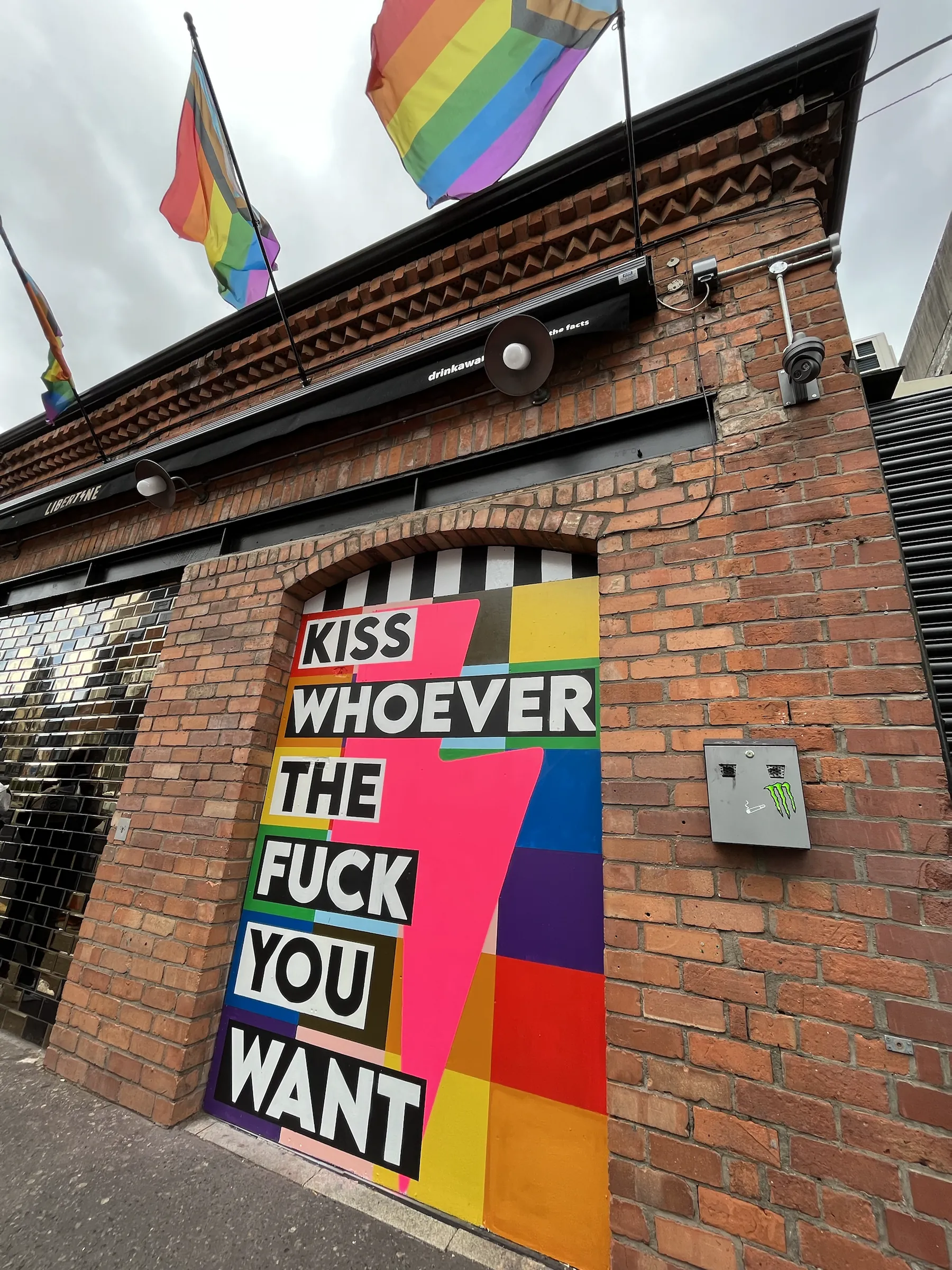 A painted doorway on a red-brick bar under rainbow flags, reading “Kiss whoever the fuck you want” in black and white blocks over bright colour bands and a pink lightning bolt.