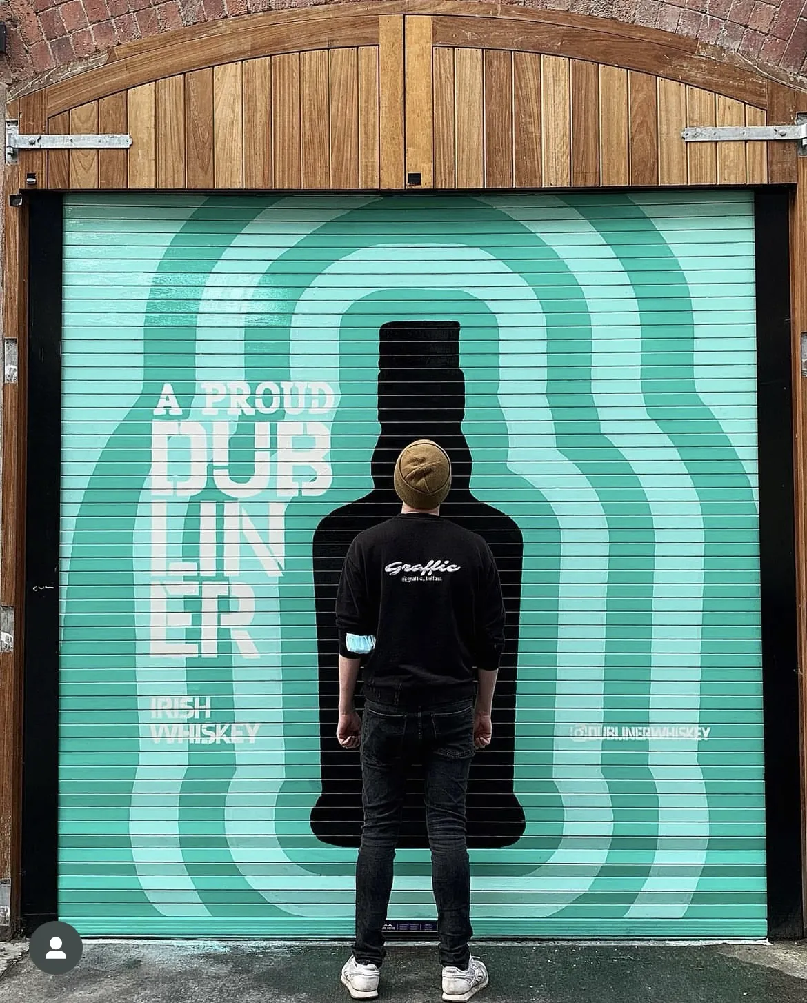 A person in a Graffic jacket standing in front of a mint-green roller shutter painted with rippling outlines of a whiskey bottle and the words “A proud Dubliner, Irish whiskey”.