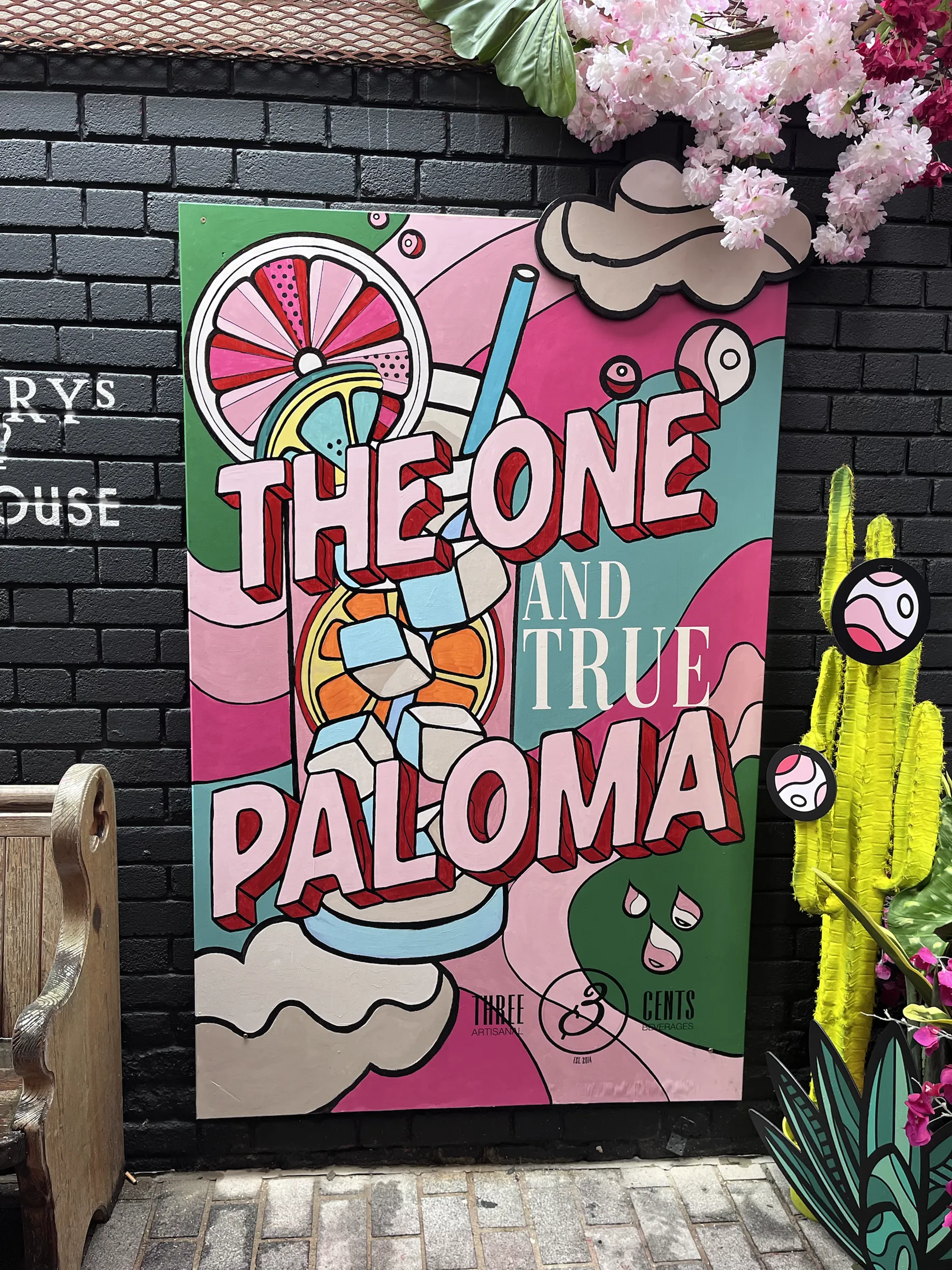 A painted panel on a black brick wall showing a giant grapefruit cocktail and the words “The one and true Paloma”, framed by pink blossom and a painted cactus.