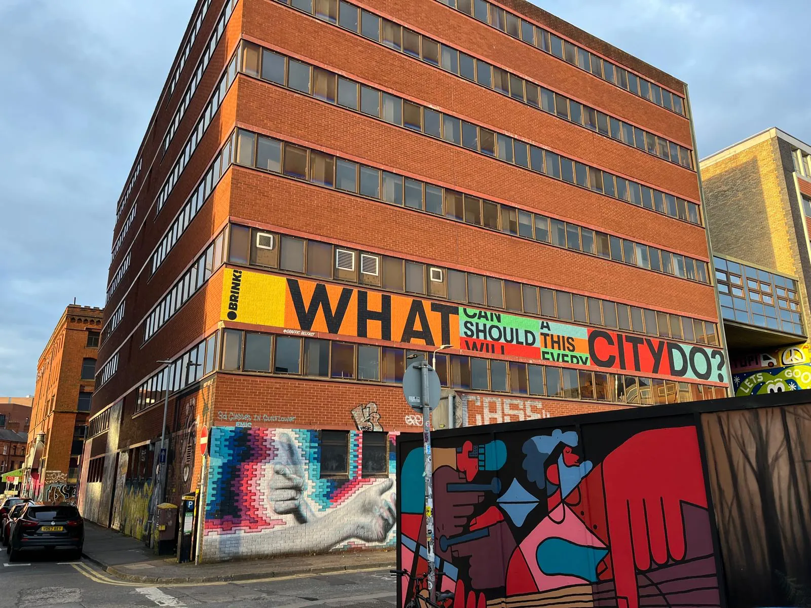 A city-centre office block with a band of large painted lettering along its first floor, above colourful murals of a giant hand and abstract figures on the street-level hoarding.