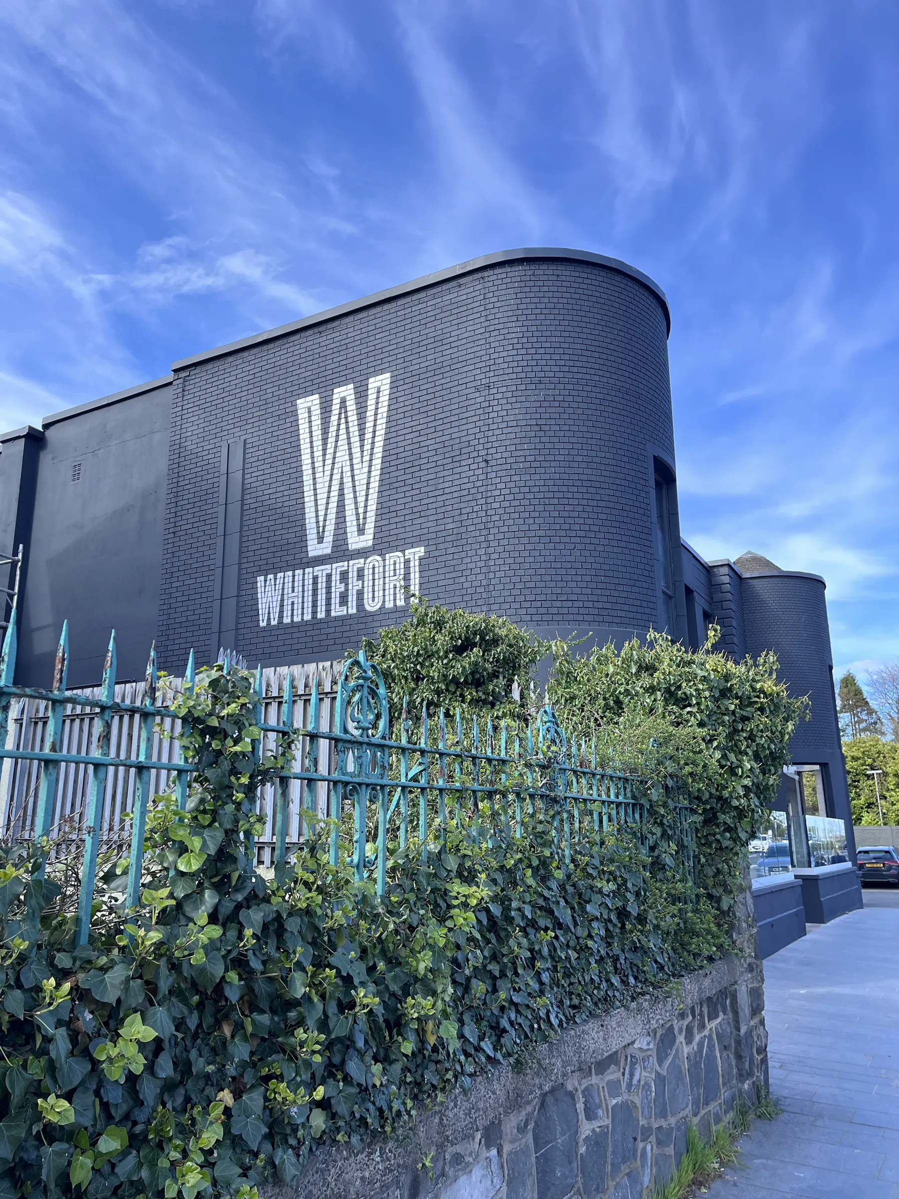 A large white “W” and the name “Whitefort” painted on the dark brick of a curved building, behind ivy-covered railings.