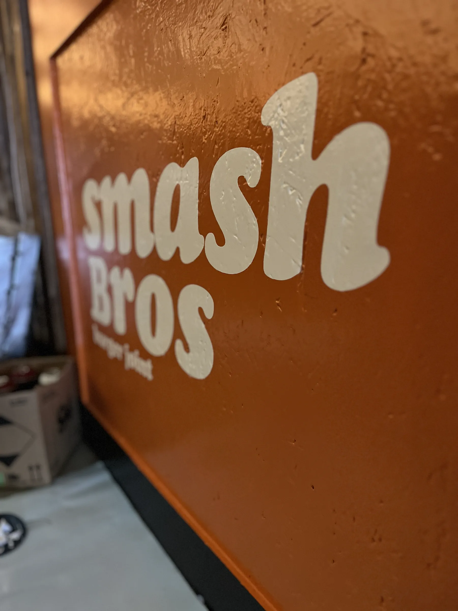 Cream lettering reading “Smash Bros, burger joint” painted on a textured orange board.