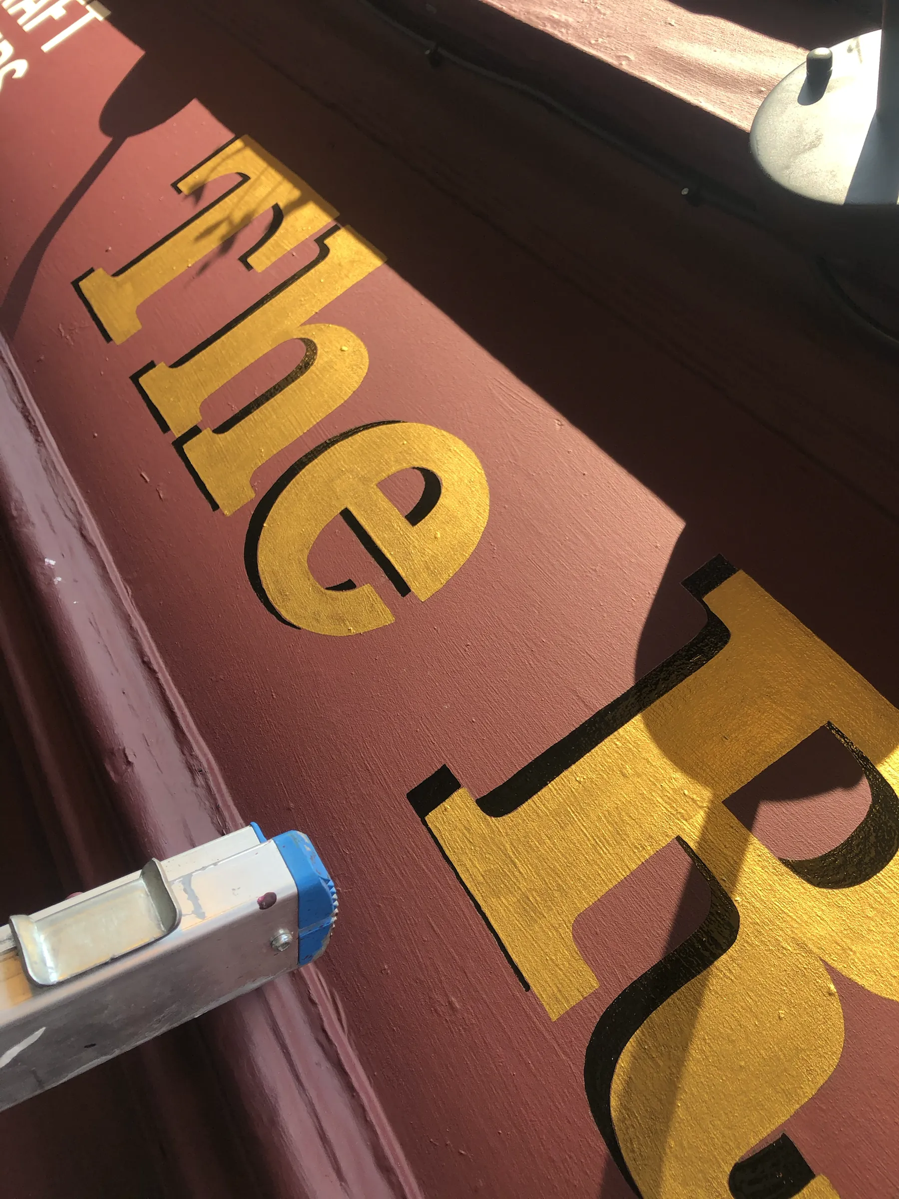 Yellow lettering with a black drop shadow being painted on a maroon pub fascia, the top of a stepladder in the foreground.