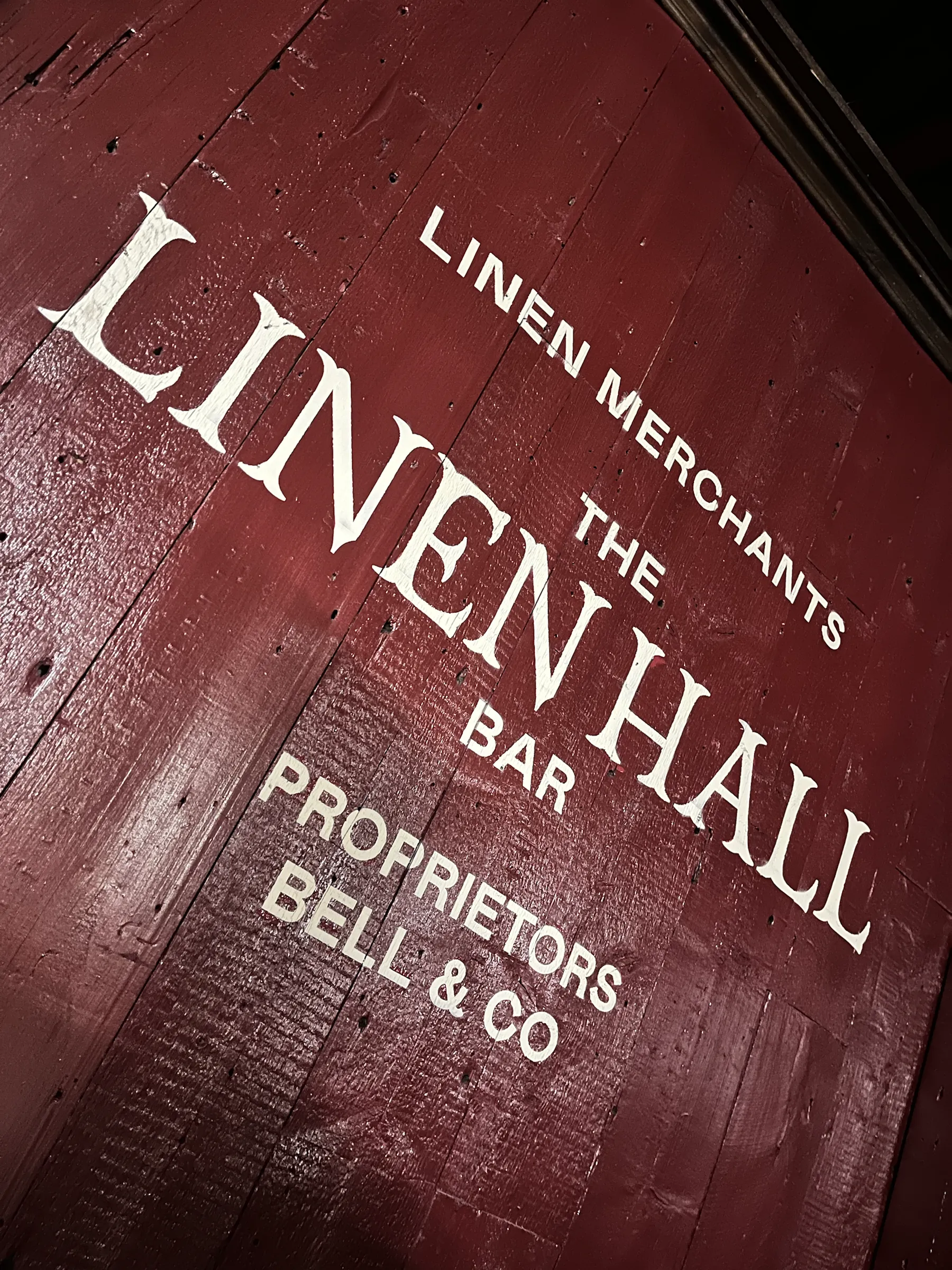 White lettering on a dark red timber wall reading “Linen Merchants, The Linen Hall Bar, Proprietors Bell & Co”.