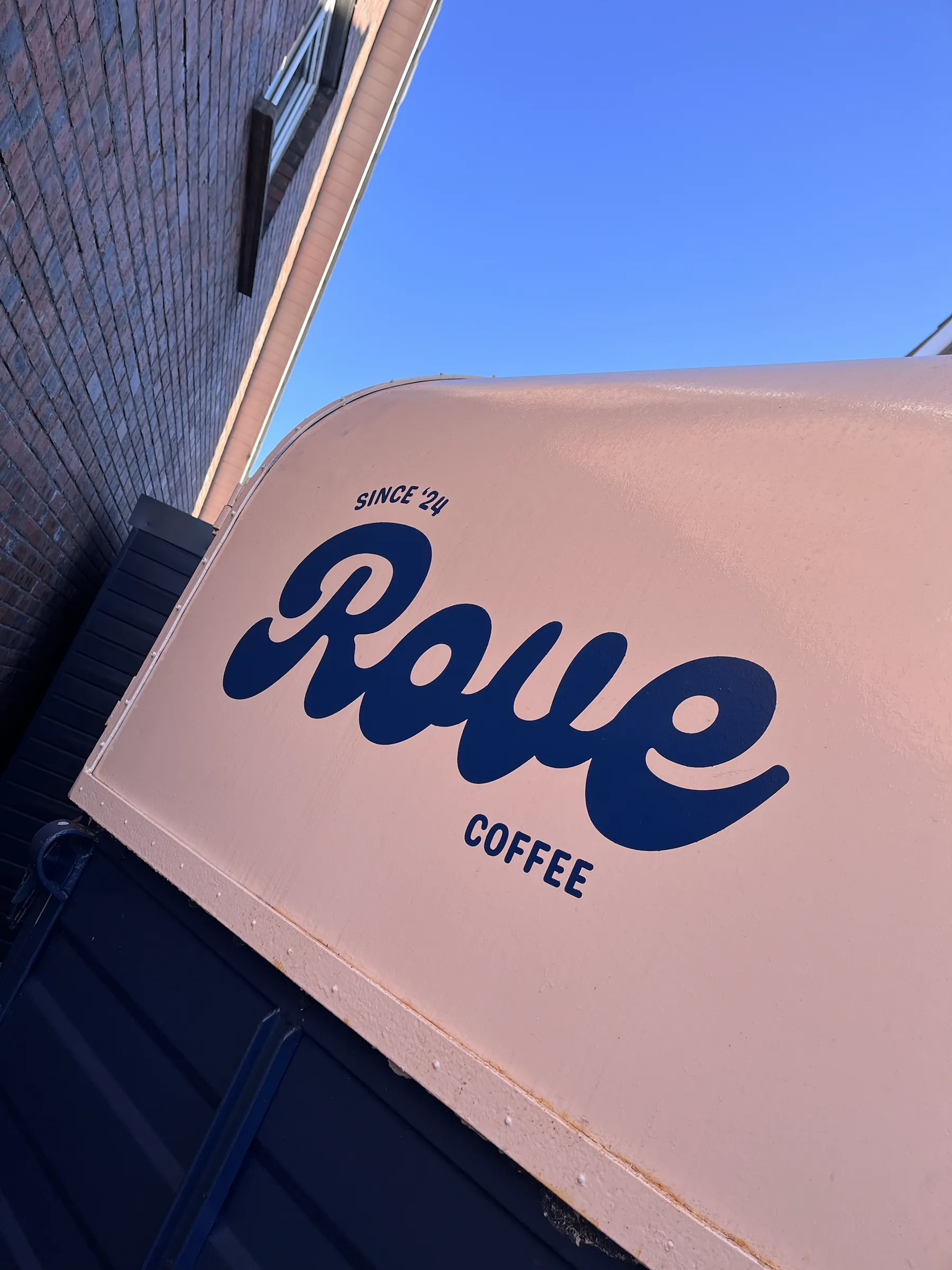 Navy script lettering reading “Rove Coffee, since ’24” painted on a pale pink canopy against a blue sky.