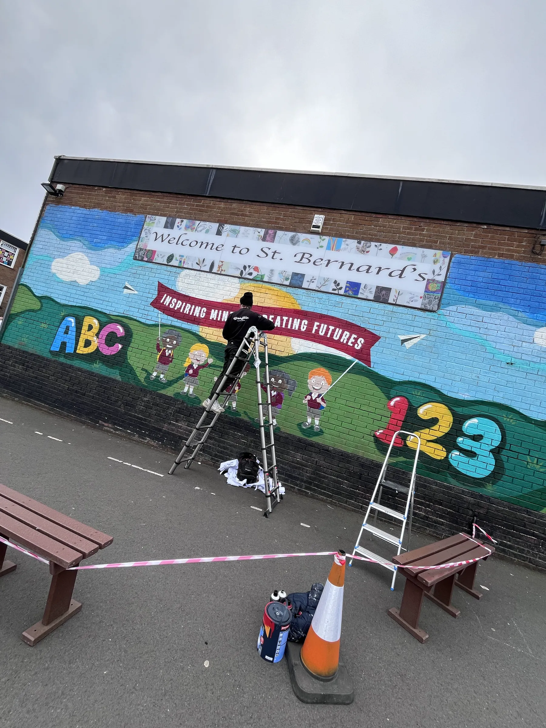 A painter on a ladder at work on the playground wall under “Welcome to St. Bernard’s”, with ABC and 123 painted either side.