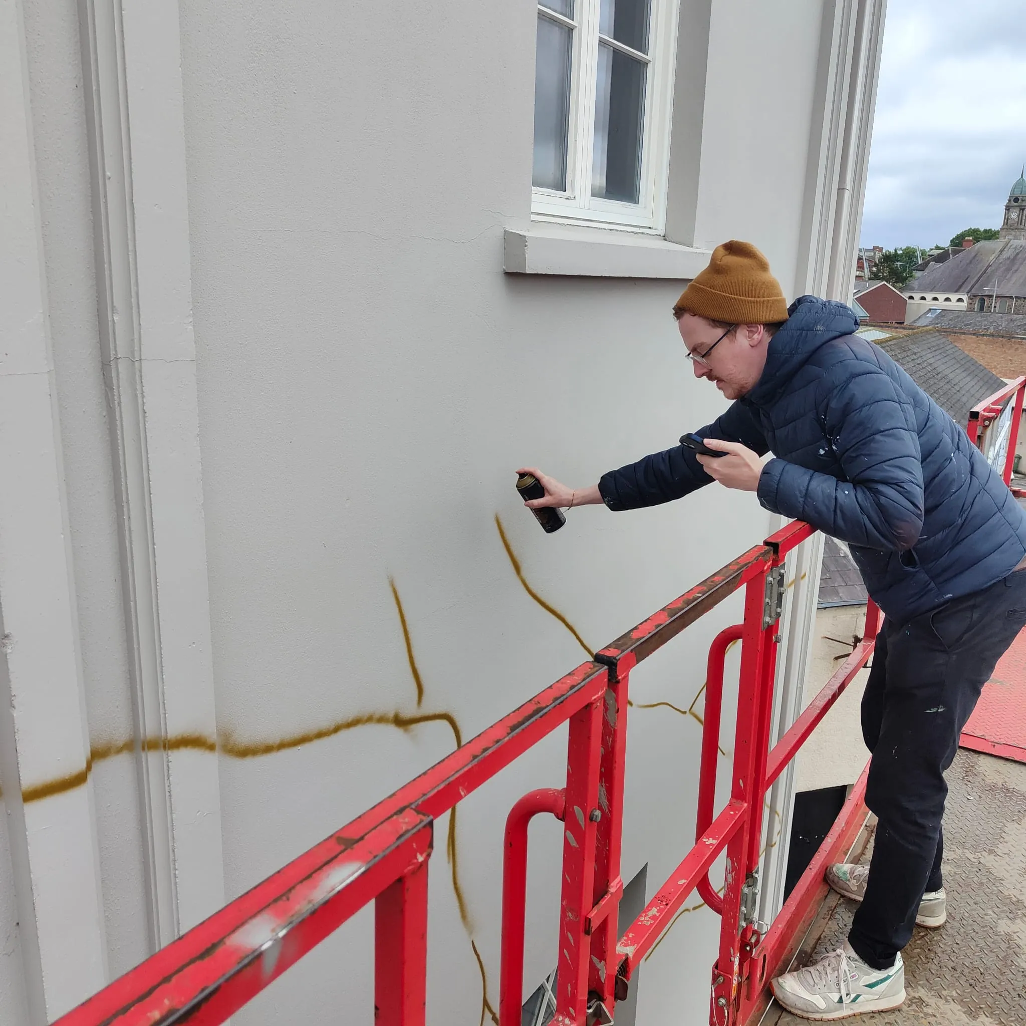 A painter in a beanie spraying gold outlines on The Lark’s white wall from a cherry picker.