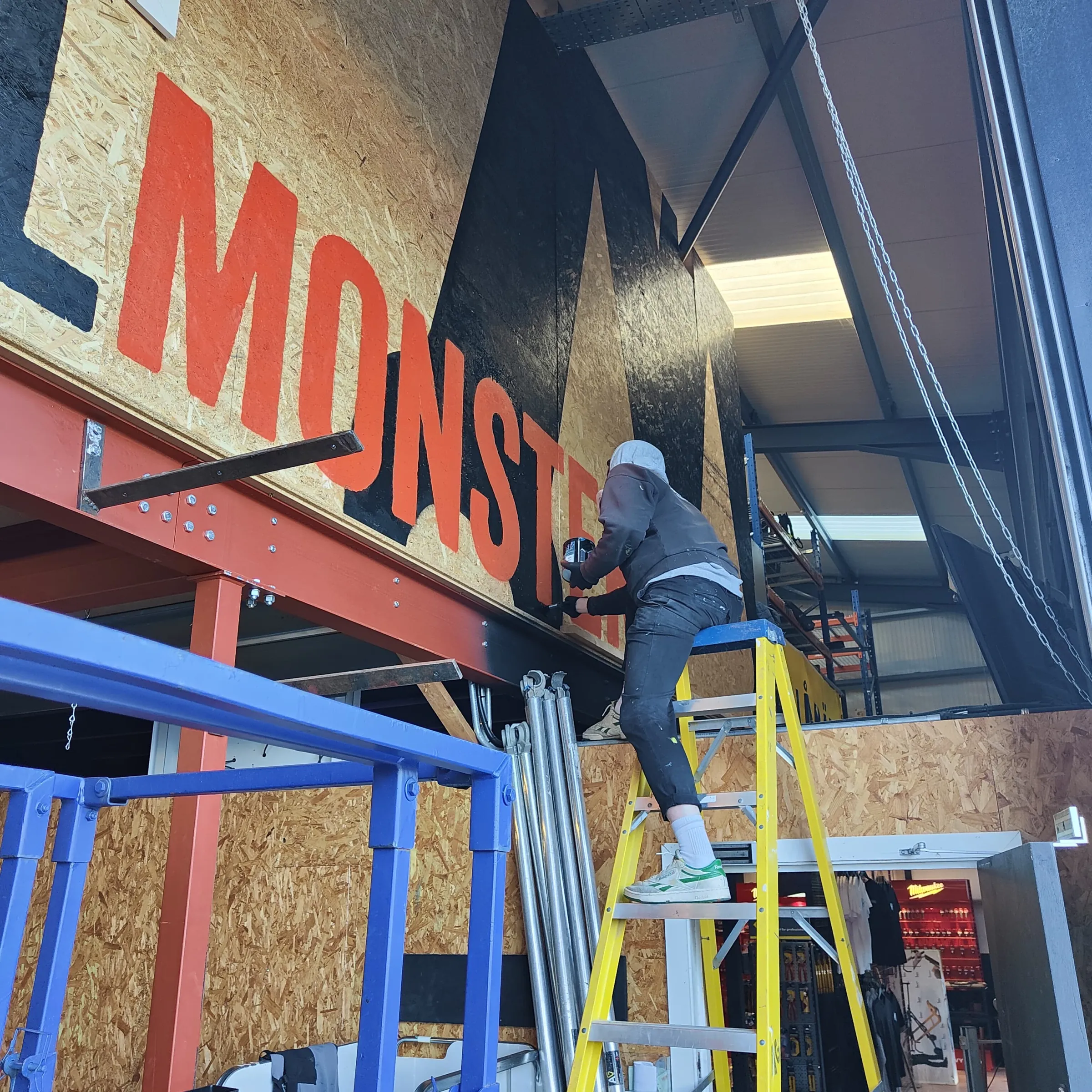 A painter on a ladder lettering “Tool Monster” in orange on the plywood wall above a mezzanine.
