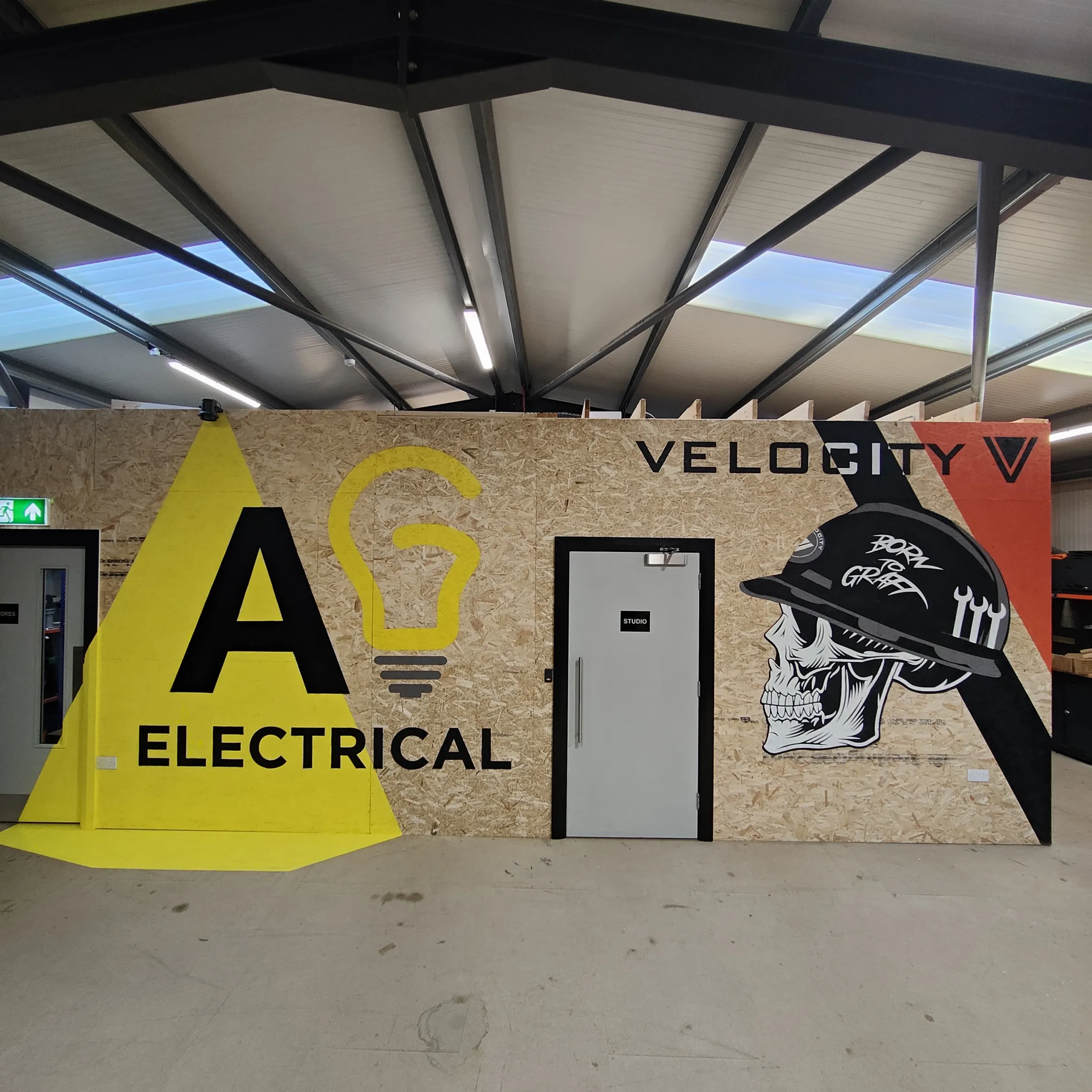 The finished workshop wall: AG Electrical’s yellow lightbulb logo, a Velocity skull in a hard hat and the Velocity name across the plywood.