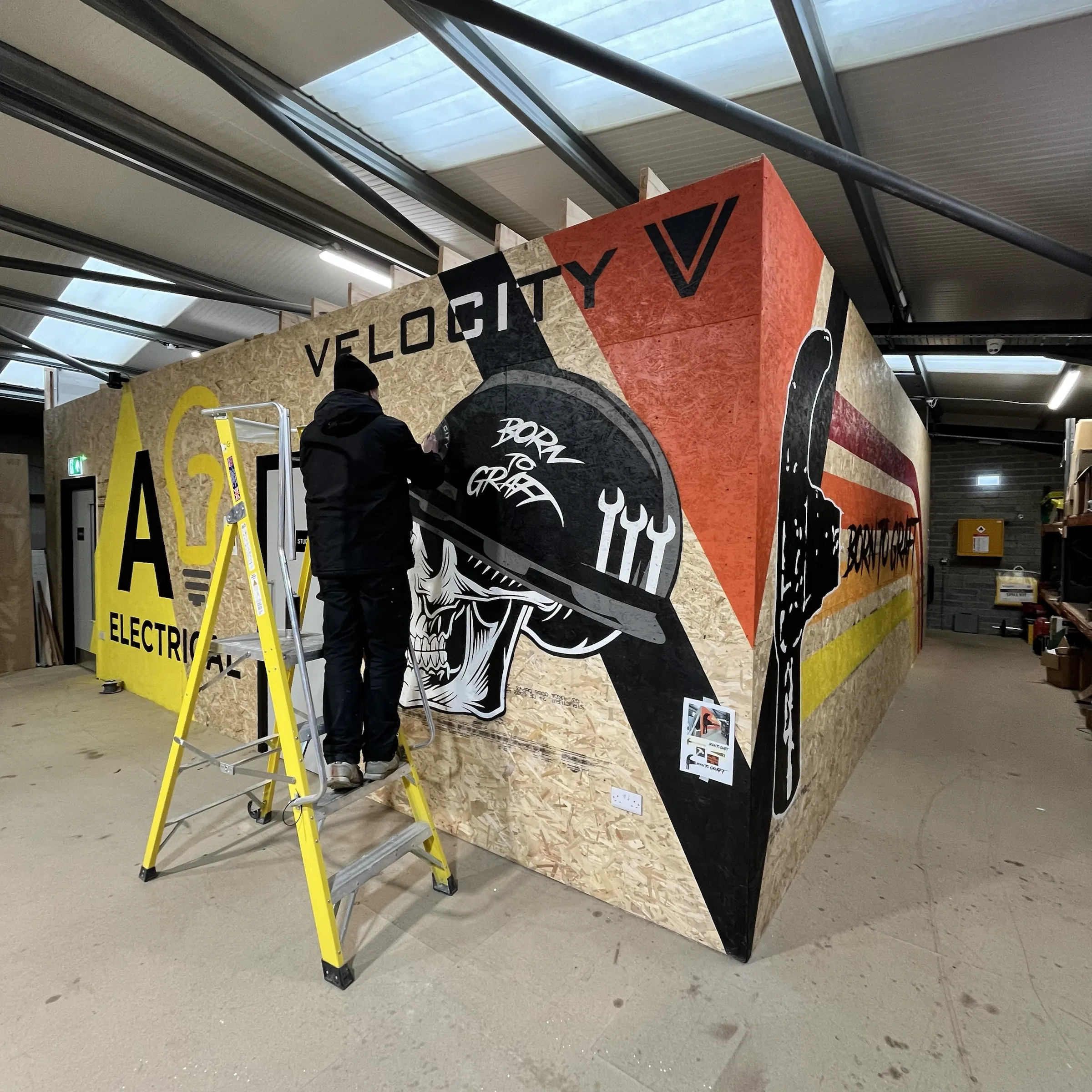 A painter on a stepladder at work on the Velocity skull, with AG Electrical’s yellow panel further along the wall.