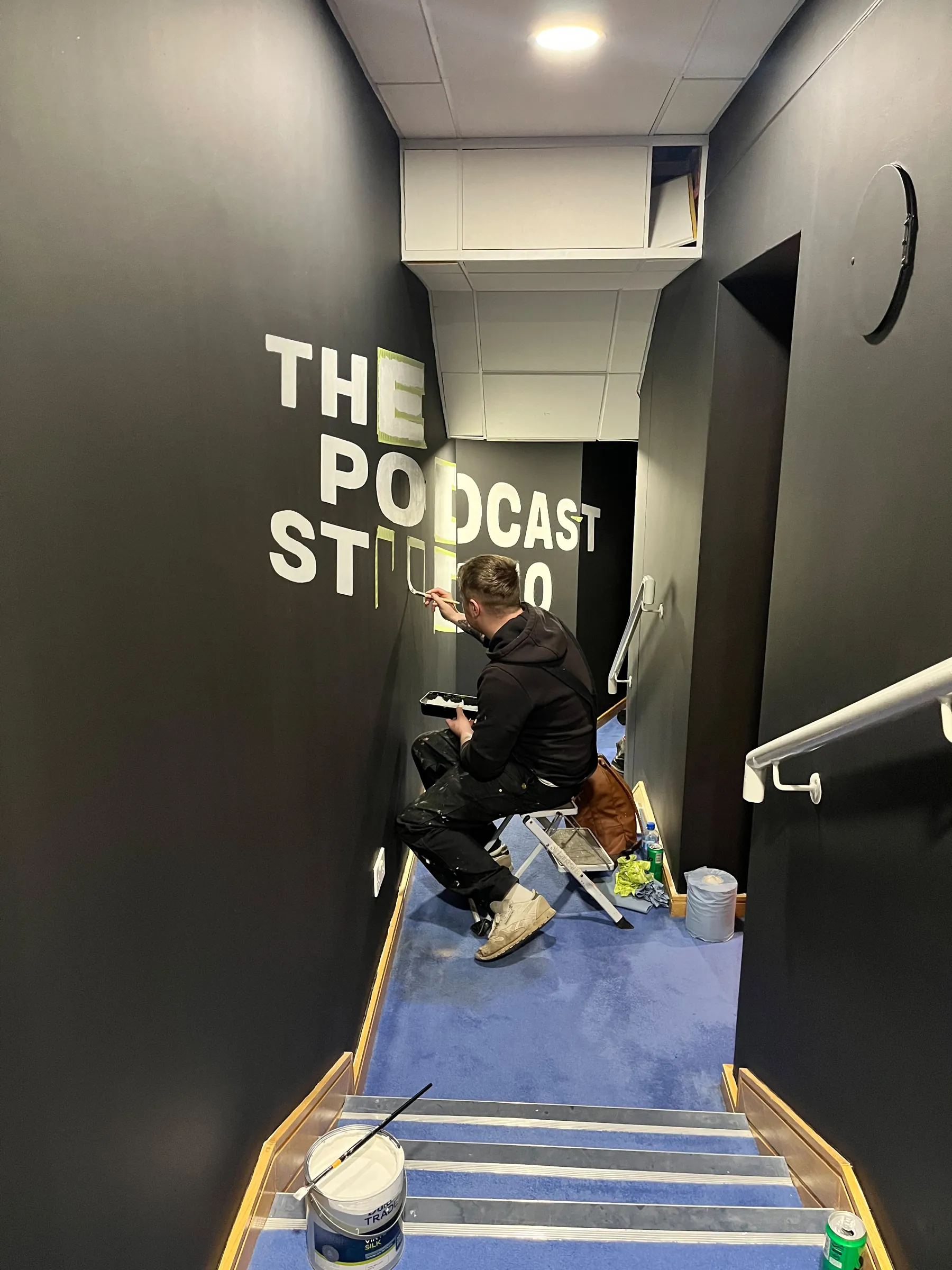 A painter seated on a step stool lettering “The Podcast Studio” on the black stairwell wall.