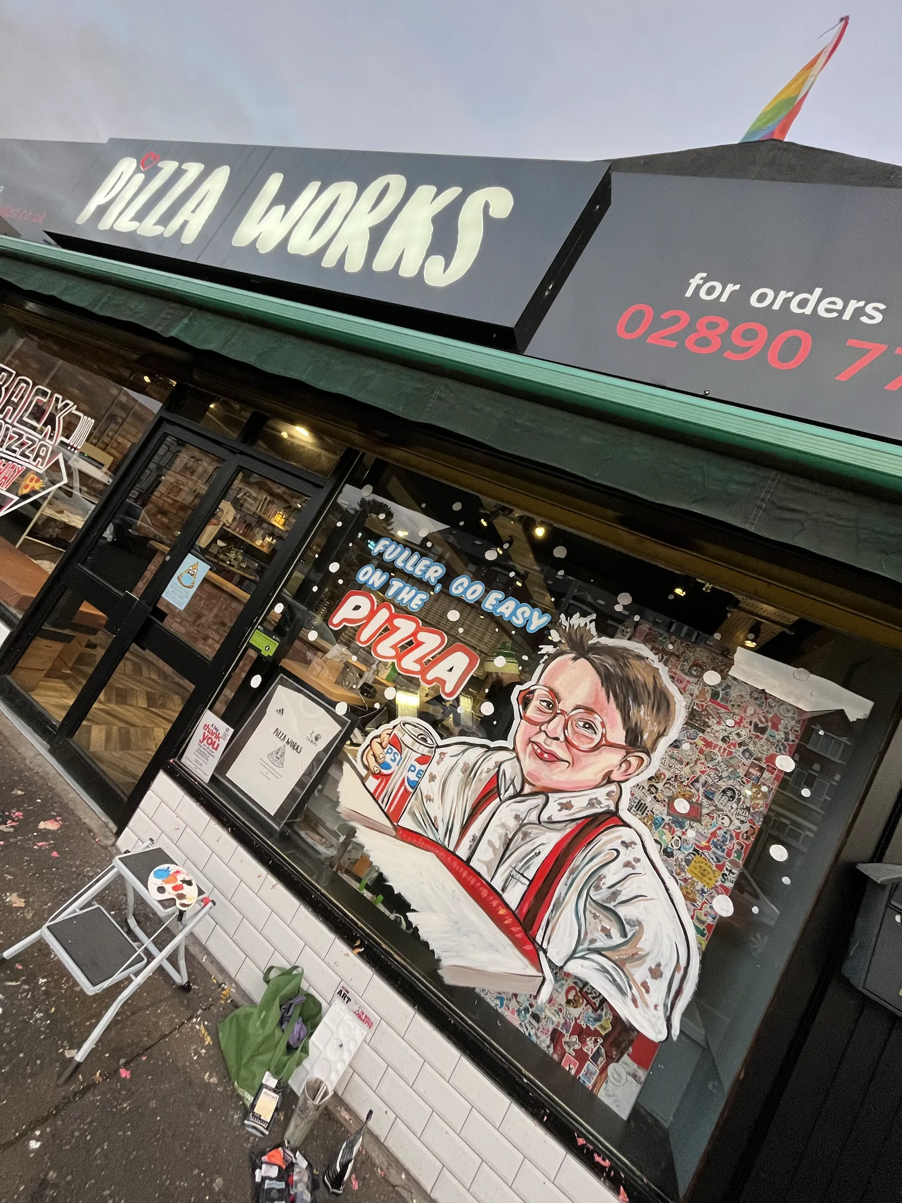The Pizza Works shopfront mid-paint, its window showing a boy in glasses holding a slice of pizza under the word “Pizza”, with paints and a folding chair on the pavement.