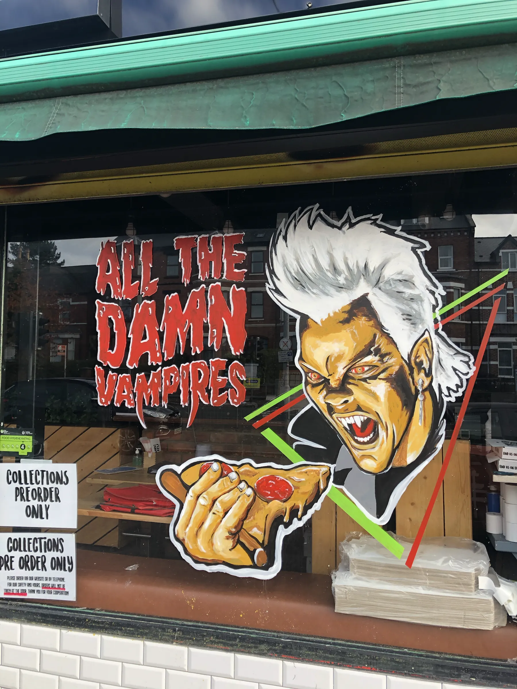 A pizza shop window painted with a snarling white-haired vampire holding a slice of pizza beside the words “All the damn vampires” in dripping red.