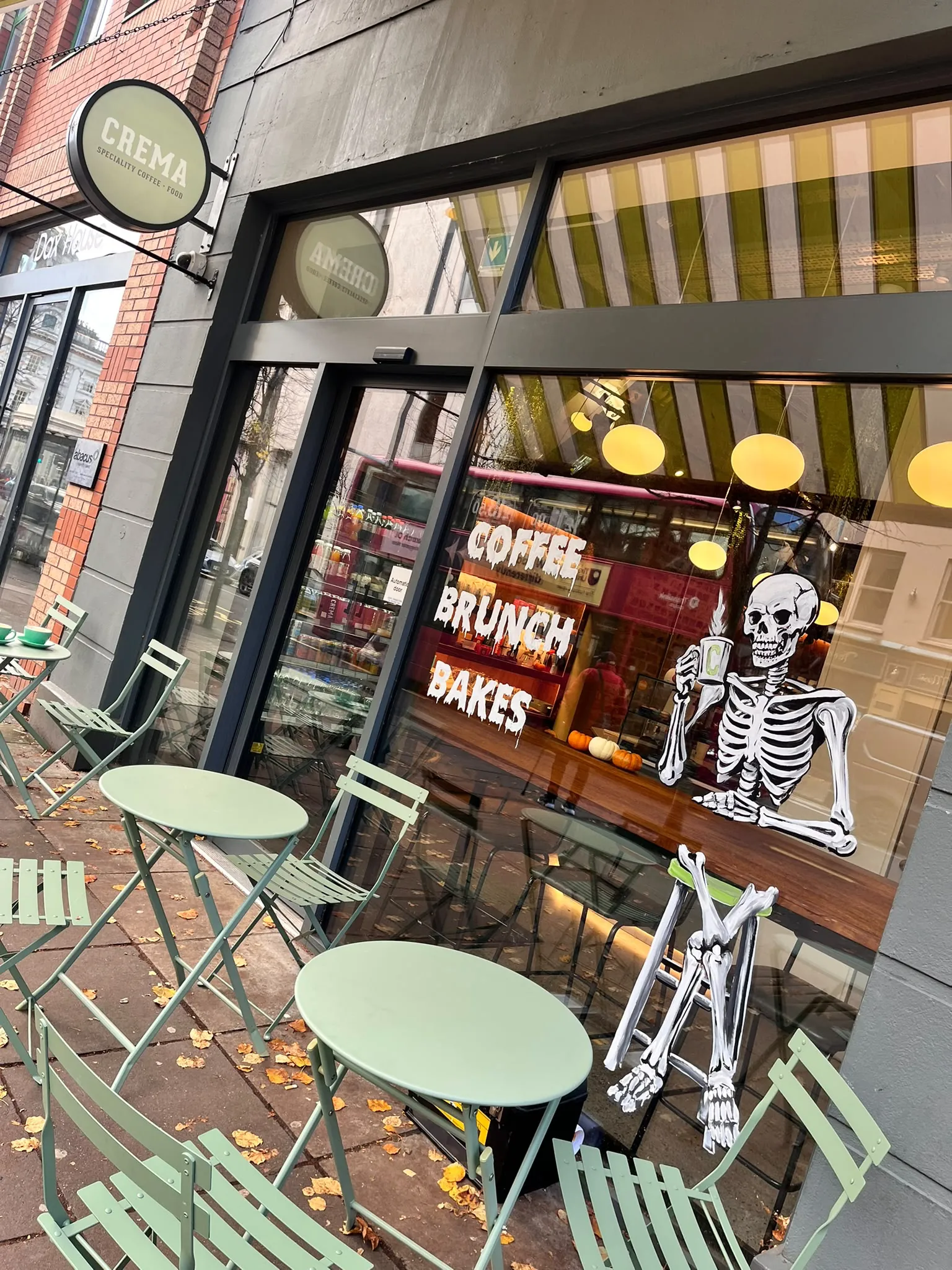 The window of Crema café painted with a grinning skeleton holding a coffee cup beside “Coffee, Brunch, Bakes” in white lettering, above green bistro tables on the pavement.