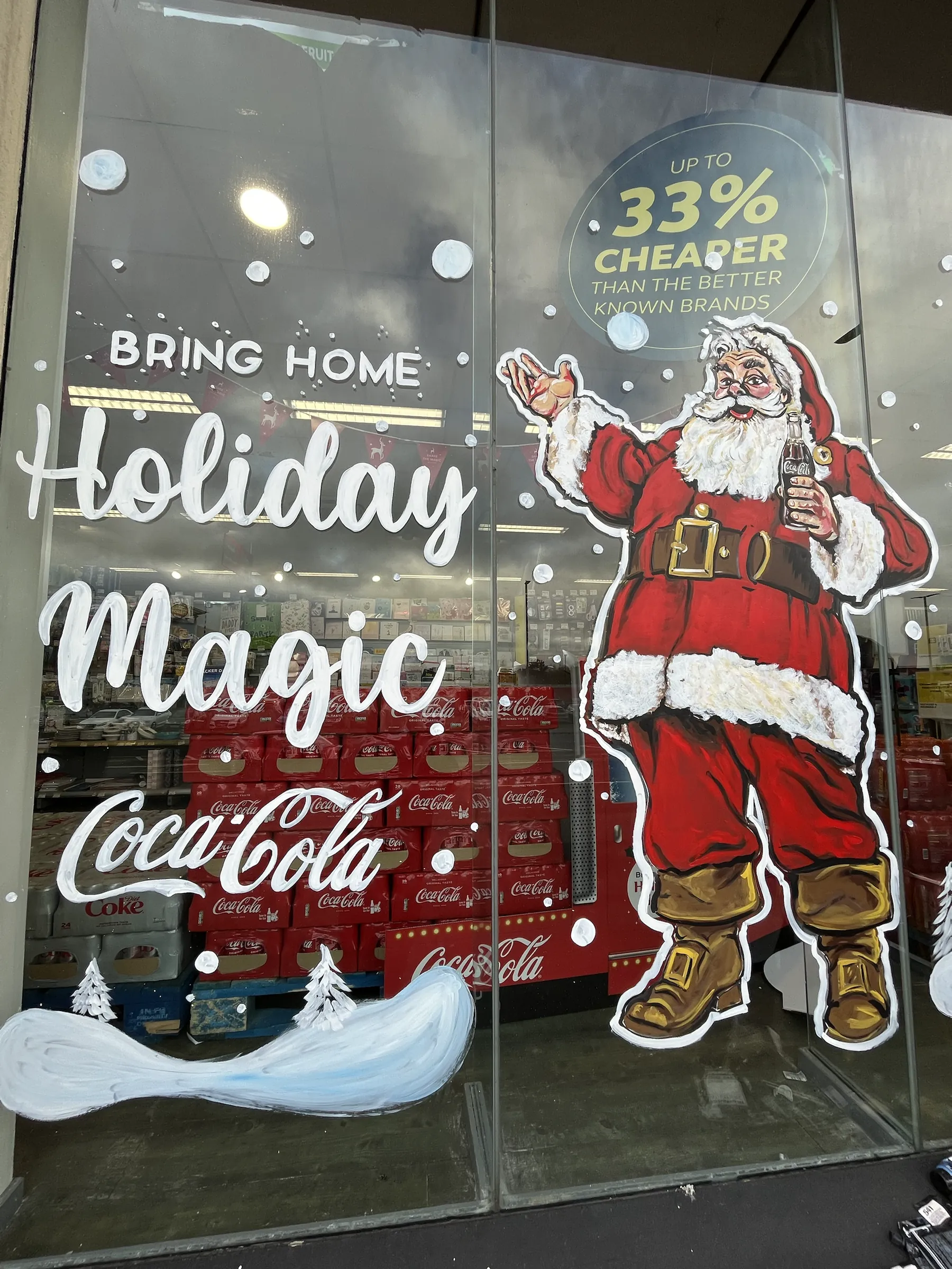 A shop window painted with a full-length Father Christmas holding a bottle of Coca-Cola beside “Bring home holiday magic” in white script, above painted snow.