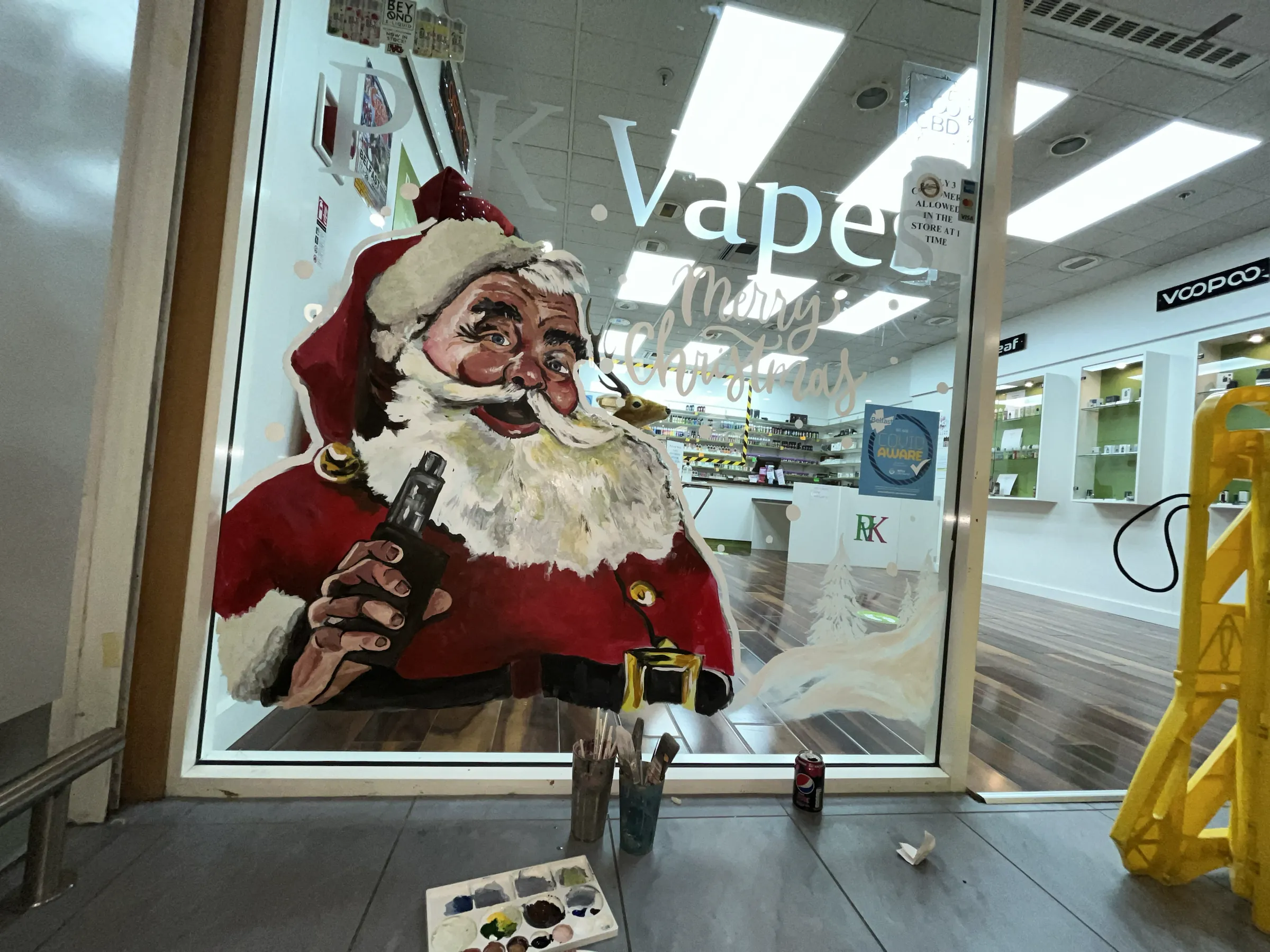 Father Christmas painted on the glass of RK Vapes in a shopping centre, with paint pots and a palette on the floor beneath the window.