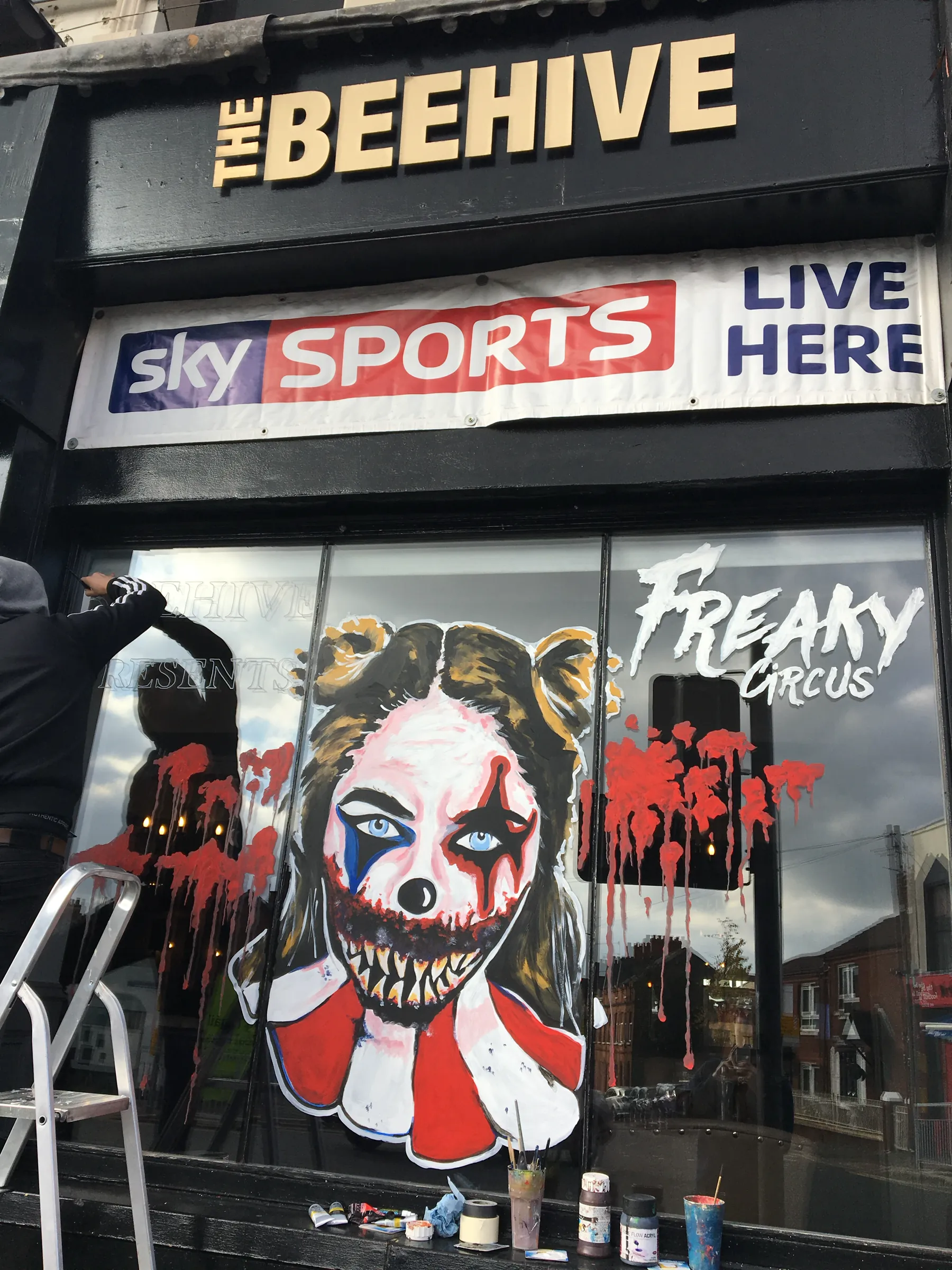 A painter on a stepladder at work on the window of The Beehive, painted with a clown-faced girl and the words “Freaky Circus” among red splatters.