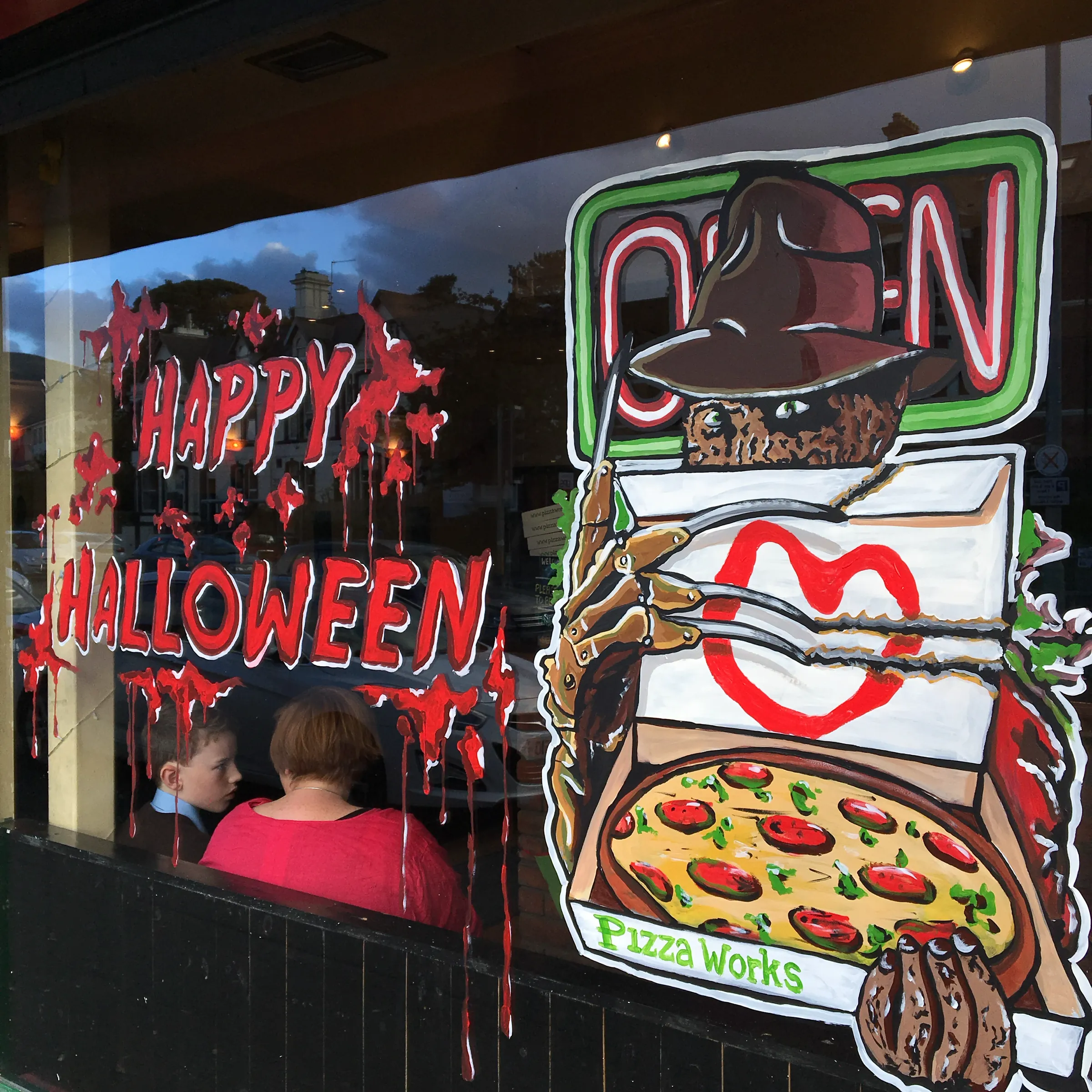 A Pizza Works window painted for Halloween, with “Happy Halloween” in dripping red beside a clawed horror villain in a fedora holding a pizza box.