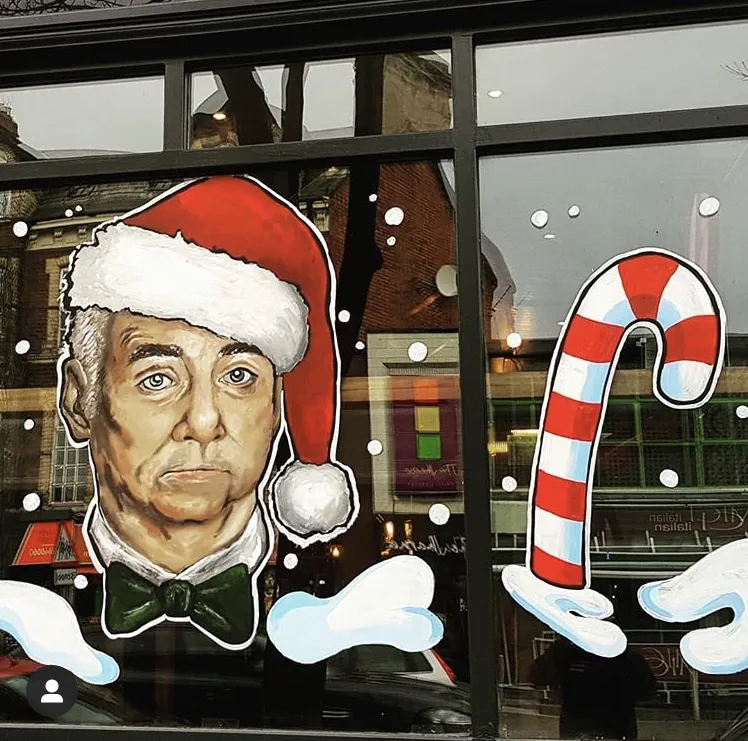 A window painted with a portrait of a grey-haired man in a Santa hat and green bow tie beside a candy cane and drifts of snow.