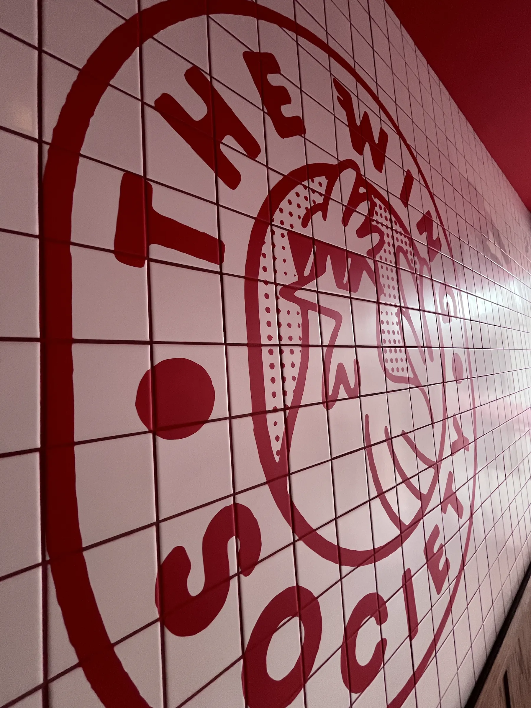 The Wing Society logo seen along the tiled wall, its red lines running across the grout.